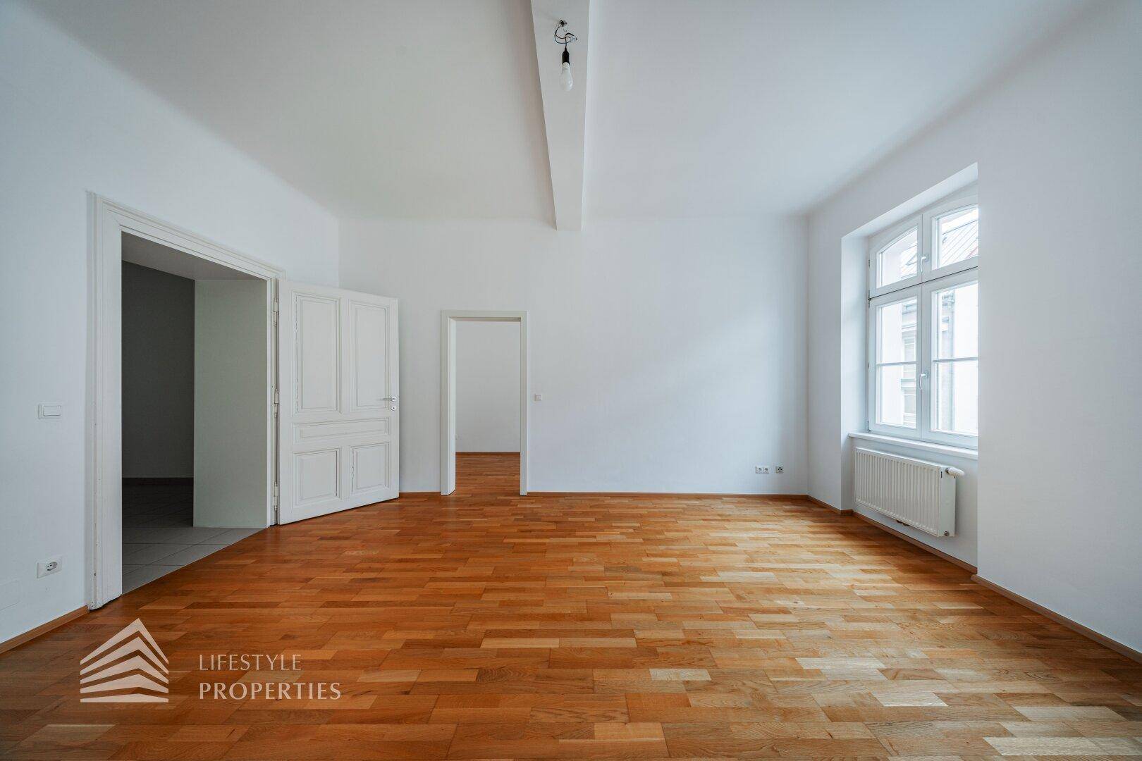 Geräumiges Zimmer mit Holzboden und weiß gestrichenen Wänden, mit Zugang zu weiteren Räumen.