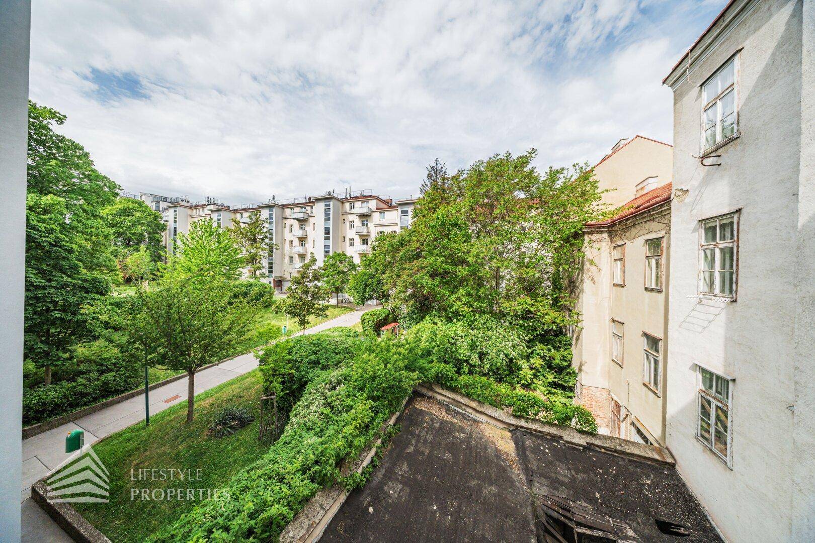 Blick aus dem Fenster auf eine grüne Parkanlage mit Bäumen und weiteren Wohngebäuden im Hintergrund.
