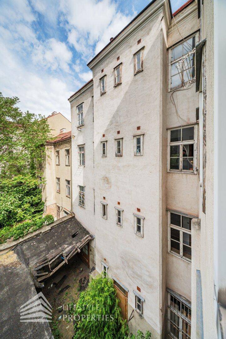 Vertikale Ansicht einer alten Gebäudeseite mit vielen Fenstern und einem verwilderten Hinterhofbereich.