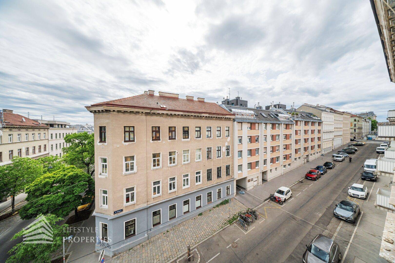 Erhöhte Ansicht einer städtischen Straße mit Gebäuden und parkenden Autos unter bewölktem Himmel.