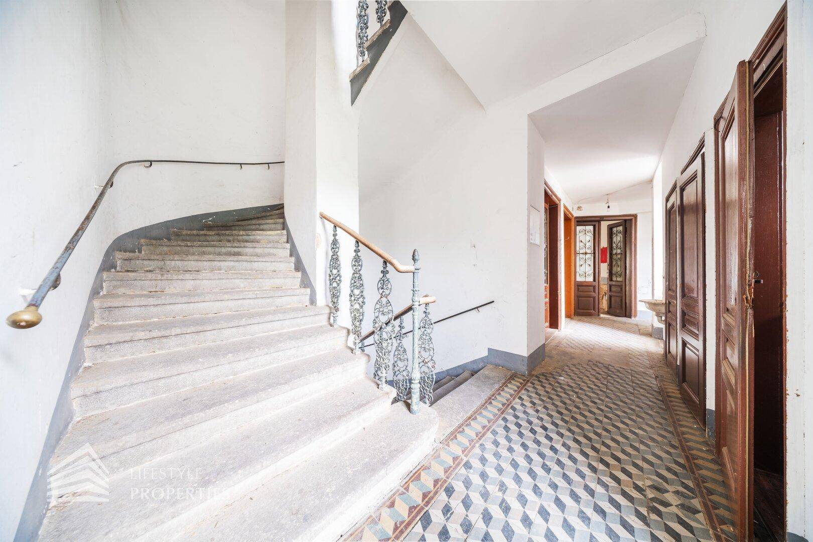 Weitläufiges Treppenhaus mit geschwungener Steintreppe, schmiedeeisernem Geländer und gemustertem Fliesenboden.