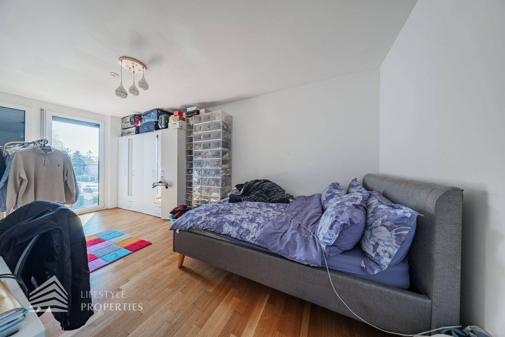 Schlafzimmer mit Holzboden, Kleiderschrank und großem Fenster mit Zugang zu einem Außenbereich.
