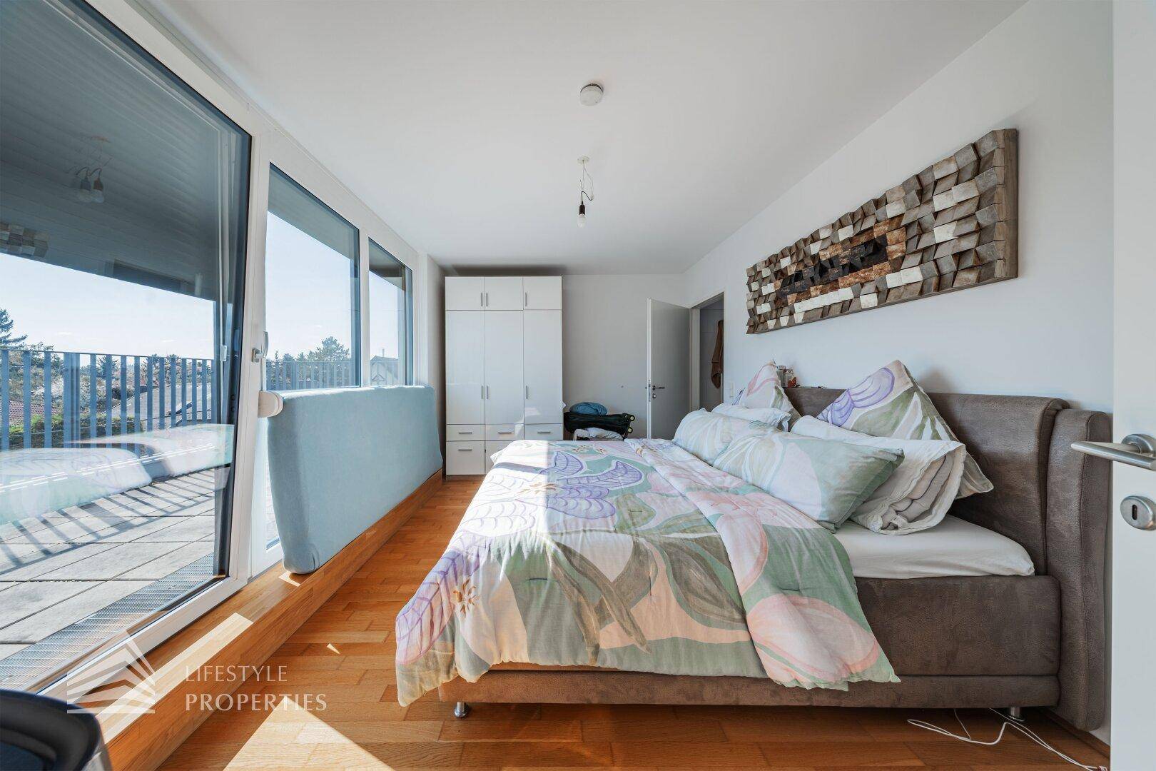 Geräumiges Schlafzimmer mit großem Bett, Holzboden und bodentiefen Fenstern zum Balkon.