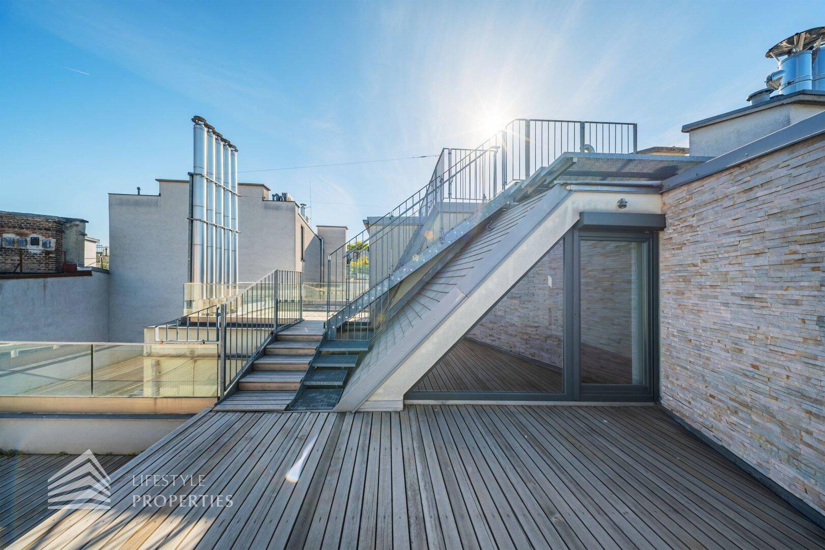 Geräumige Dachterrasse mit Holzbelag und Treppenaufgang unter strahlend blauem Himmel.
