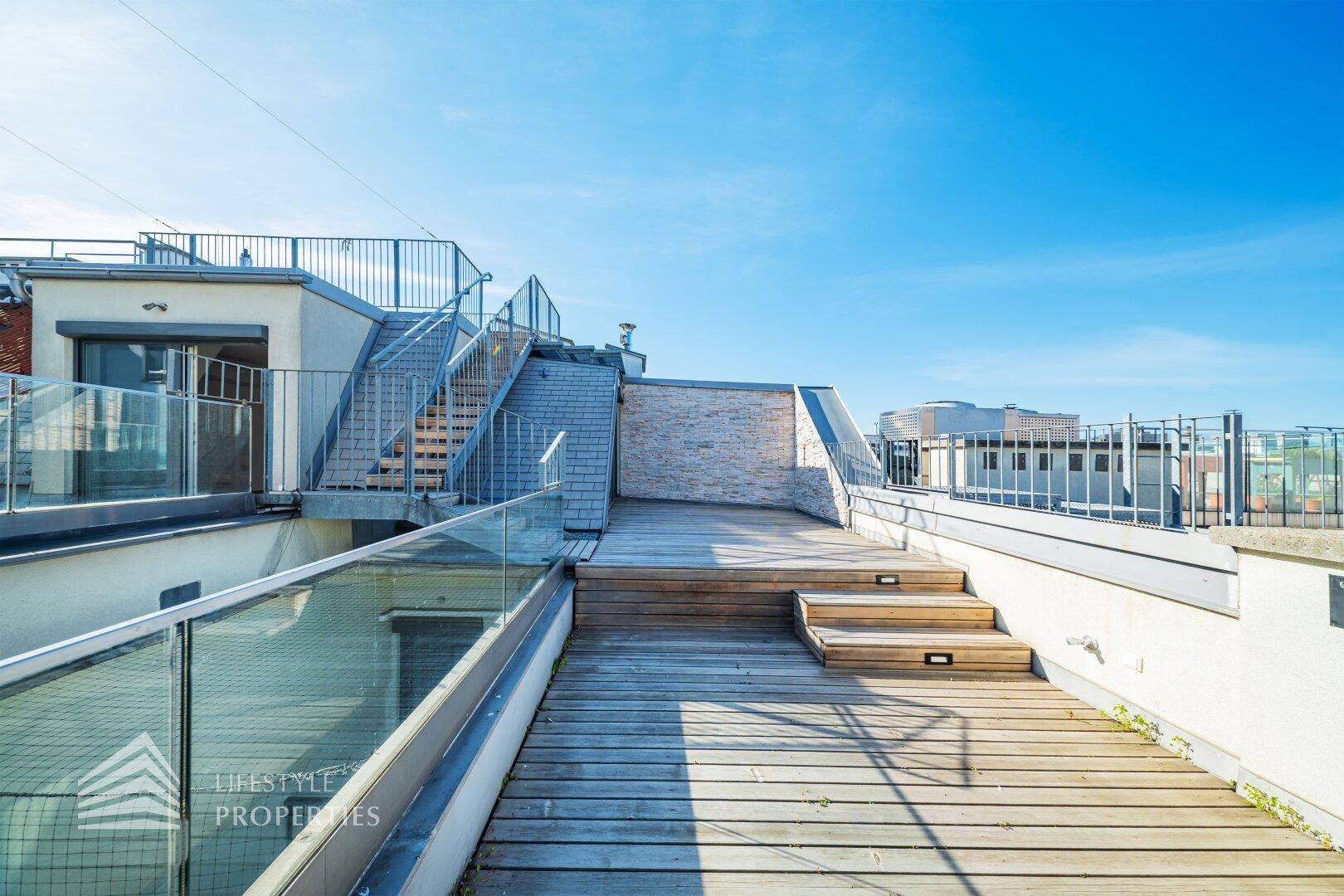 Helle Dachterrasse mit Holzbelag, Glasgeländer und Zugang zu weiteren Ebenen.