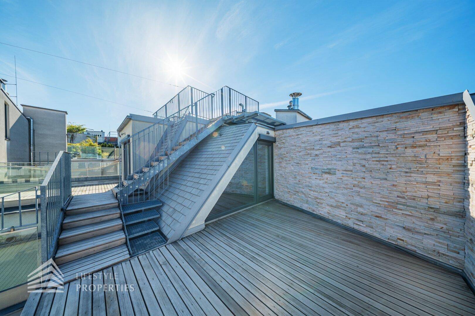 Sonnige Dachterrasse mit Holzplanken, Steinwand und modernem Treppenaufgang.