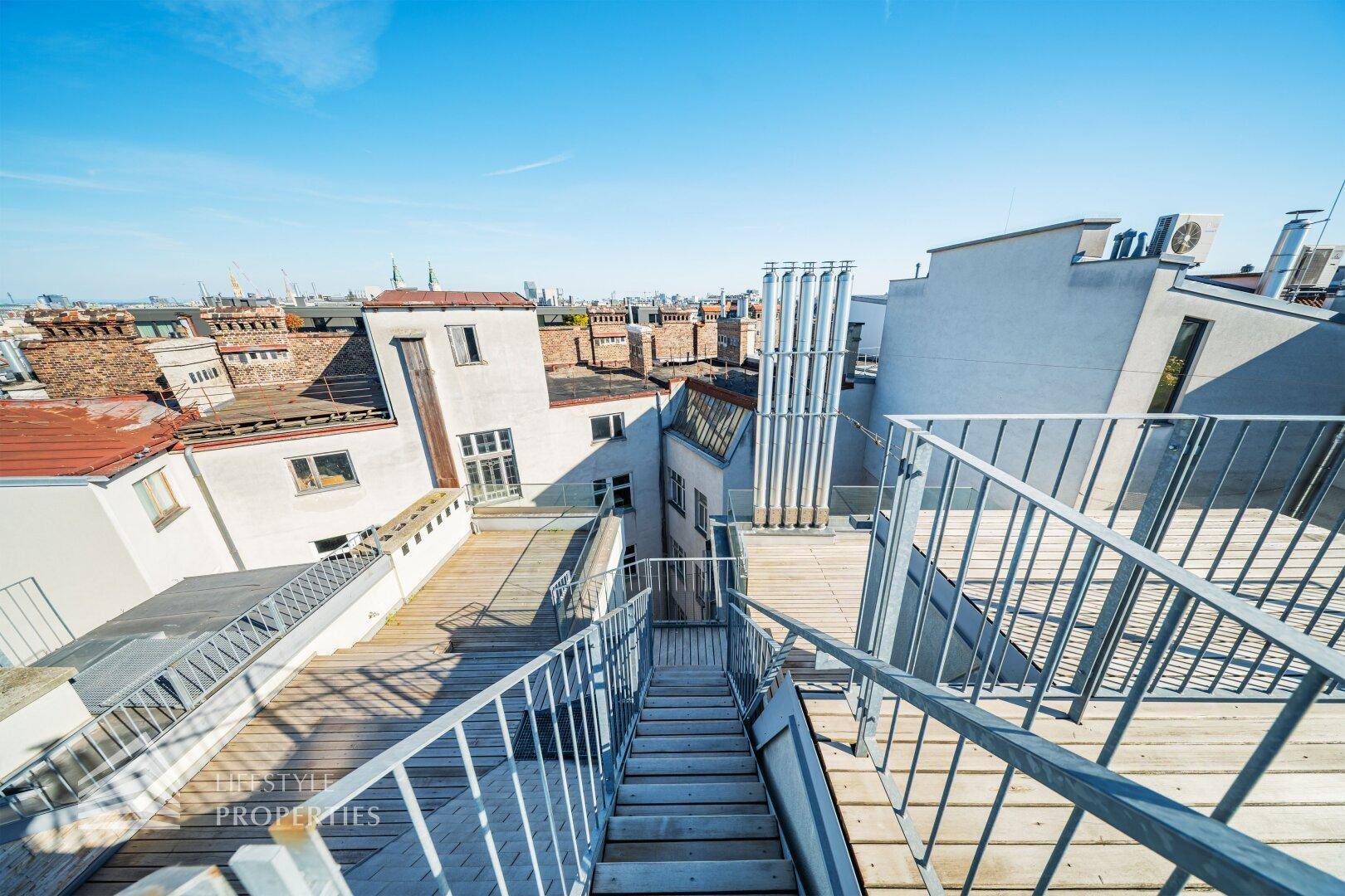 Dachterrasse mit Holzbelag und Treppenabgang, umgeben von urbaner Architektur.