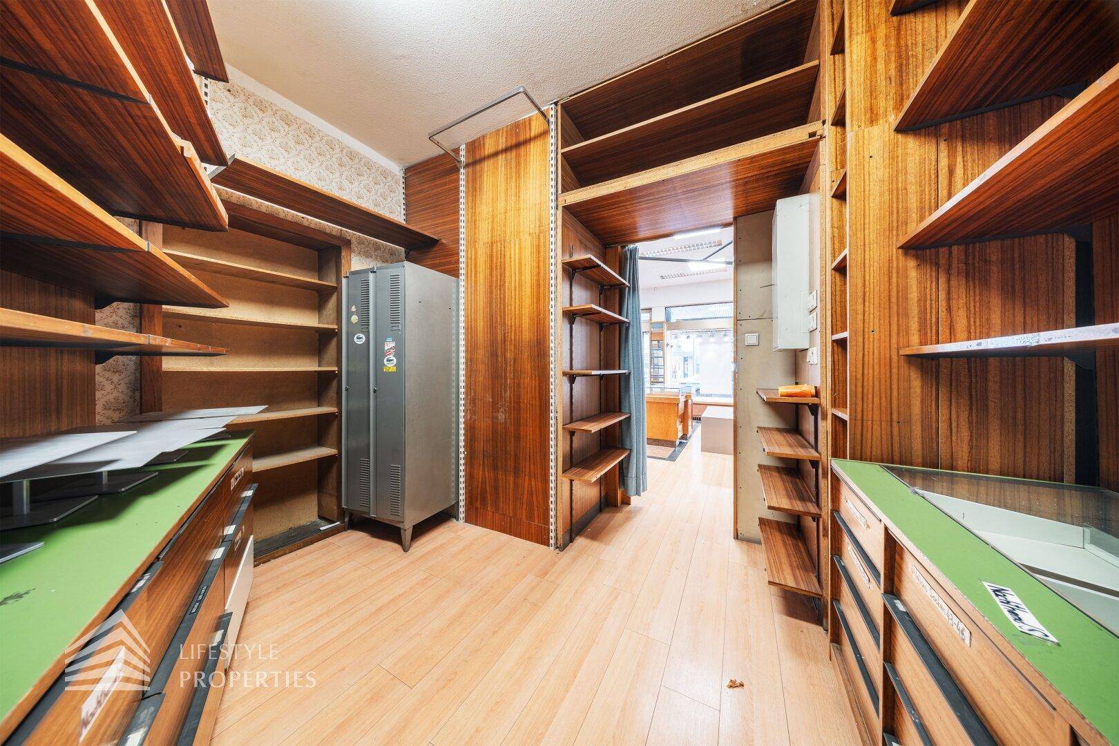 Hinterraum mit Holzregalen, einem Metallschrank und Blick in den Hauptverkaufsbereich.