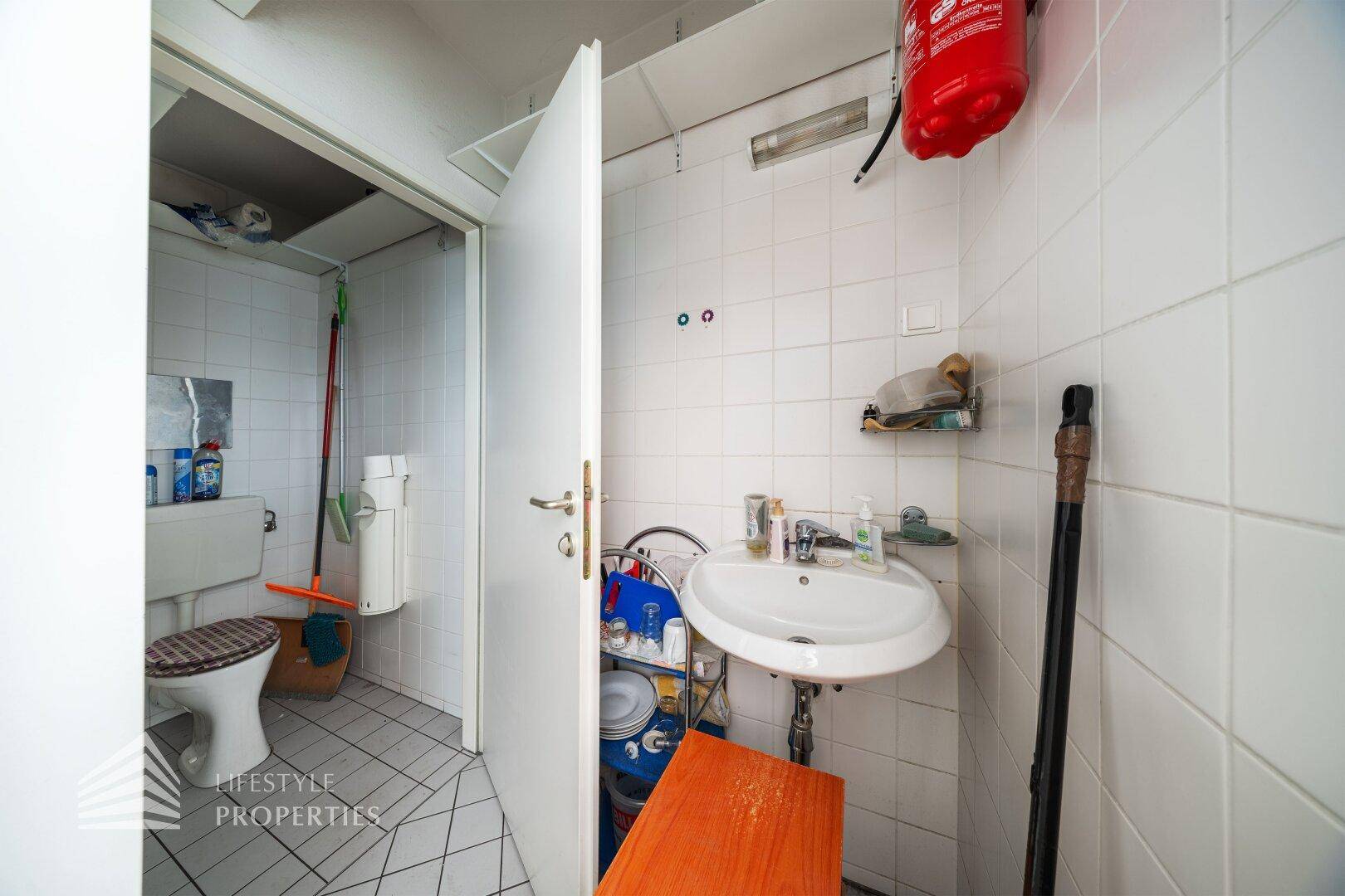 Einfache Toilette mit Waschbecken und Fliesen an Wänden und Boden, inklusive Feuerlöscher für Sicherheit.