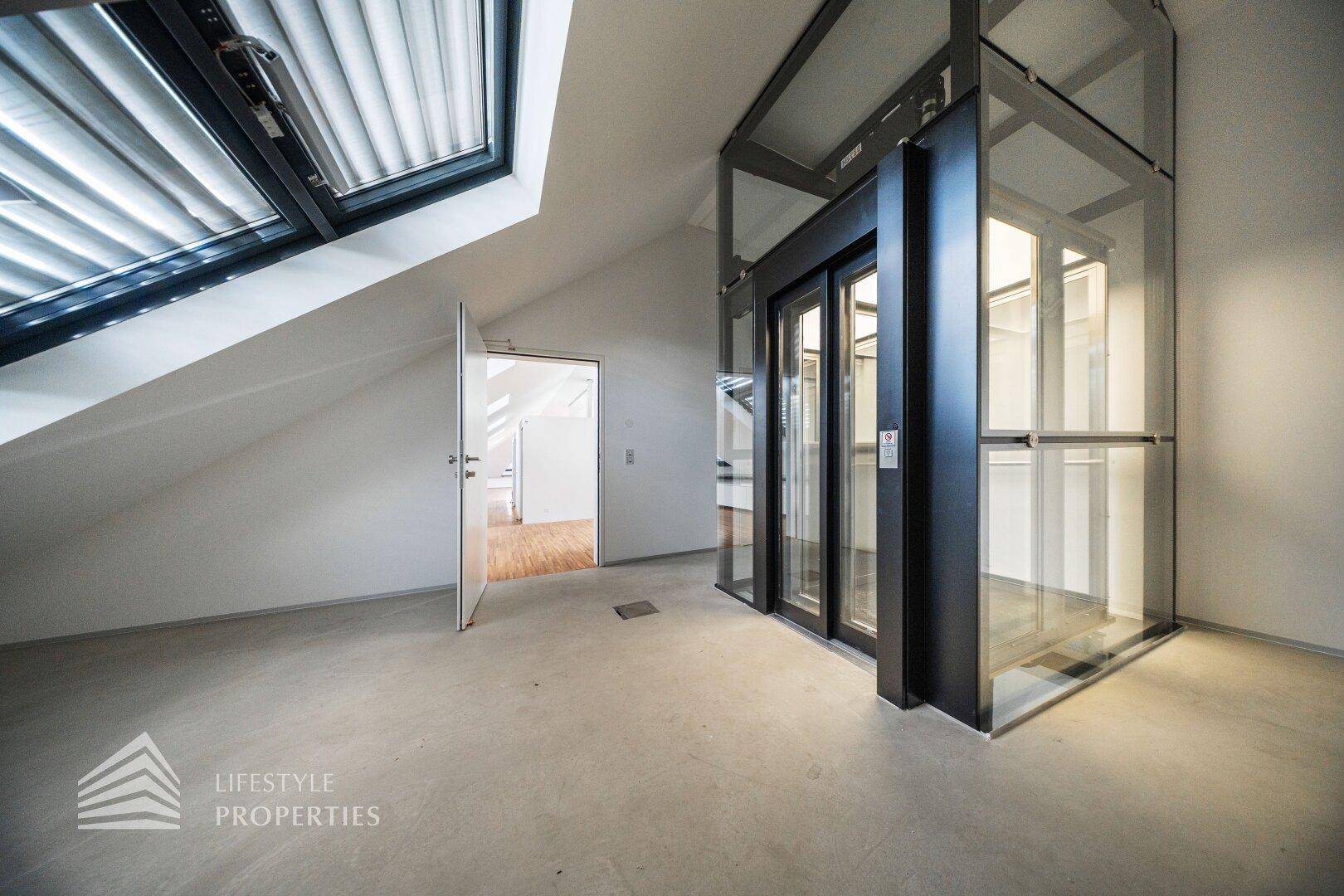 Heller Flur im Dachgeschoss mit modernem Glasaufzug, Betonboden und Zugang zu weiteren Räumen.