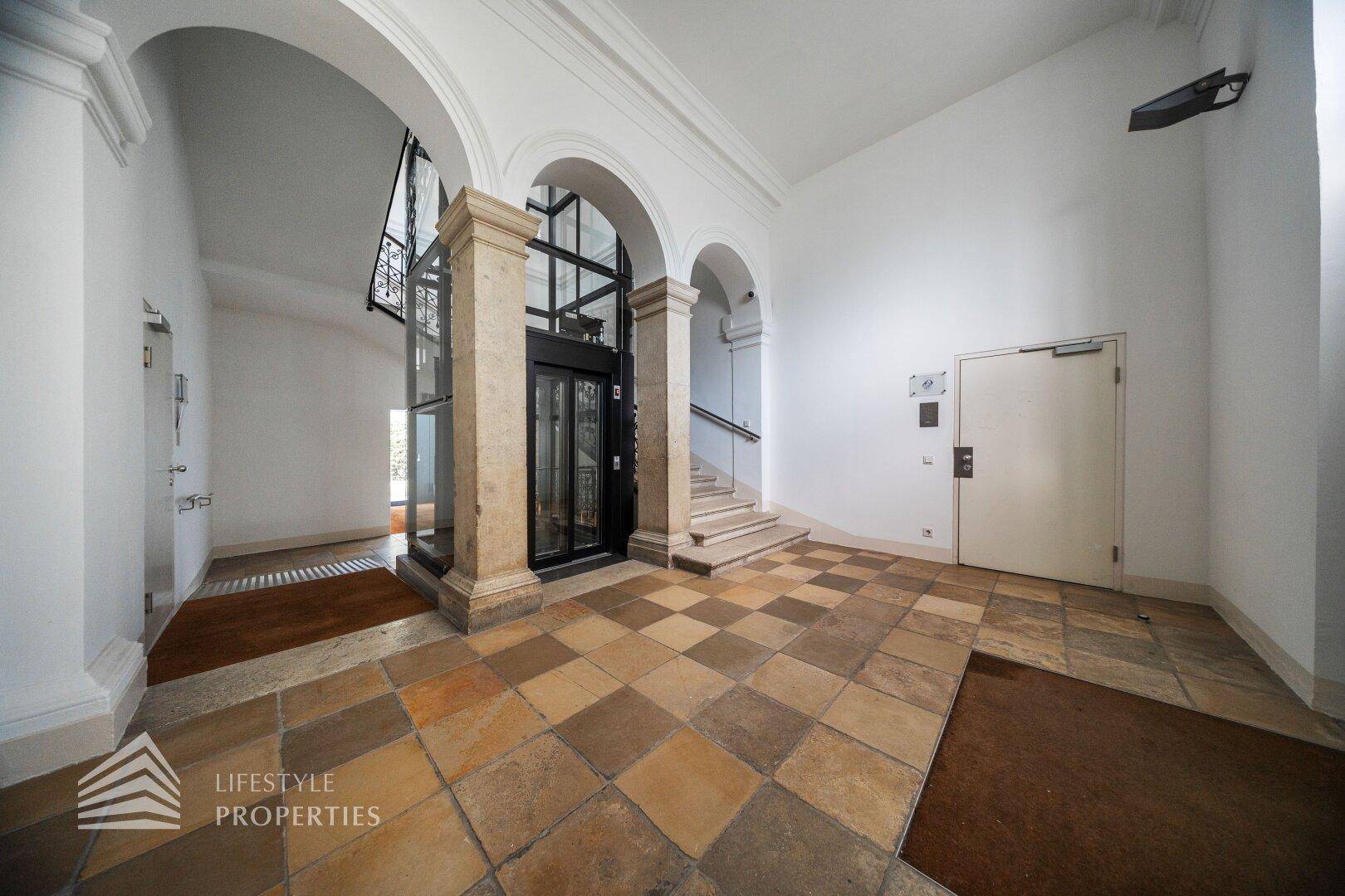 Historischer Eingangsbereich mit Säulen, elegantem Fliesenboden und einem integrierten Glasaufzug sowie Treppe.