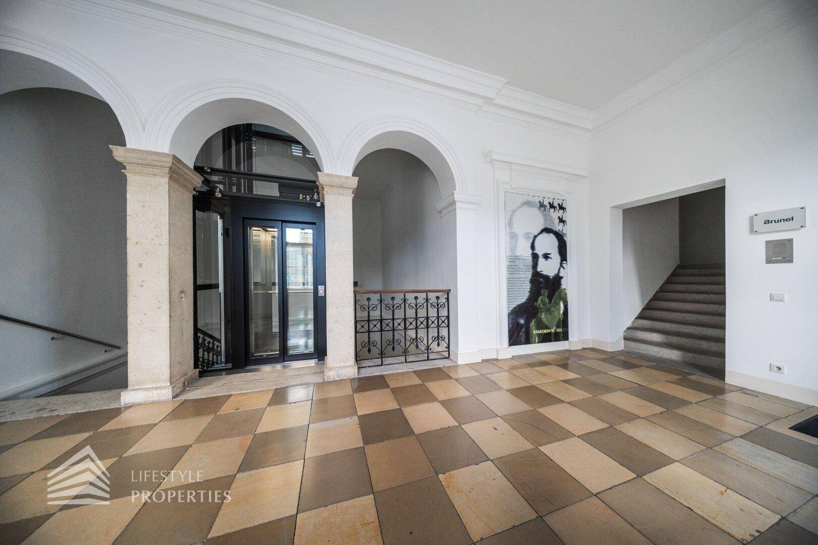 Geräumige Lobby mit historischen Architekturelementen, gemustertem Fliesenboden und Treppenaufgang neben dem Aufzug.