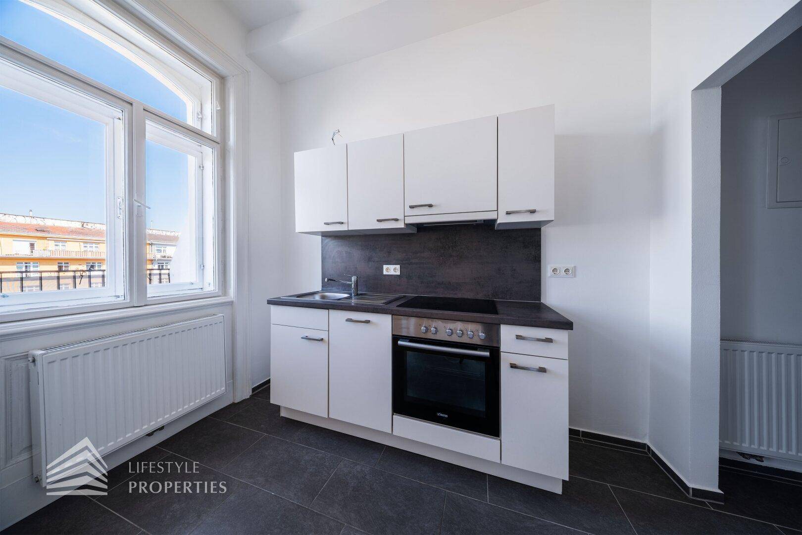 Moderne Küchenzeile mit weißen Schränken, dunkler Arbeitsplatte und integriertem Backofen, hell durch Fenster.