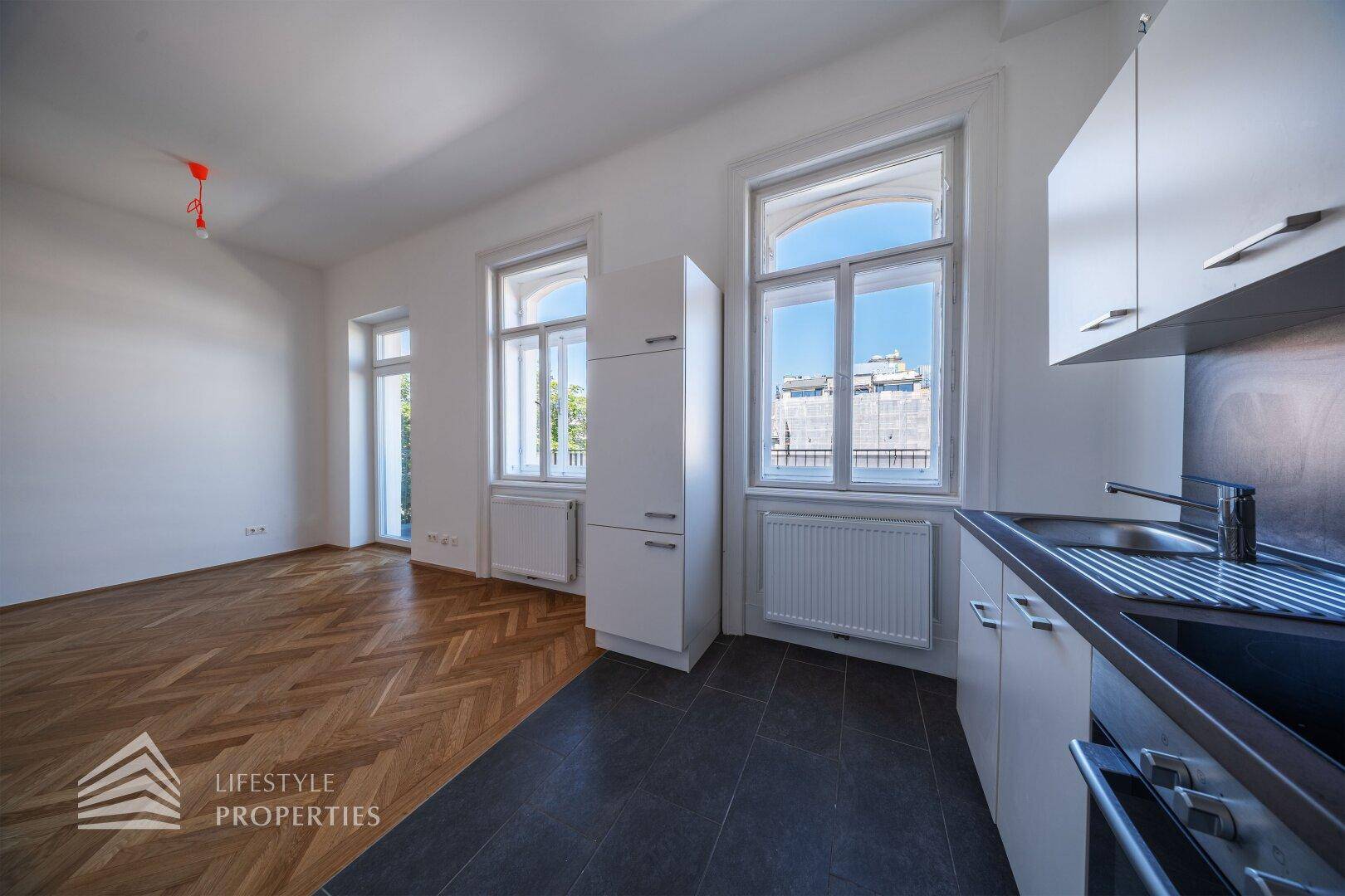 Offener Wohnbereich mit moderner Küchenzeile, Fischgrätparkett und großen Fenstern mit Stadtblick.