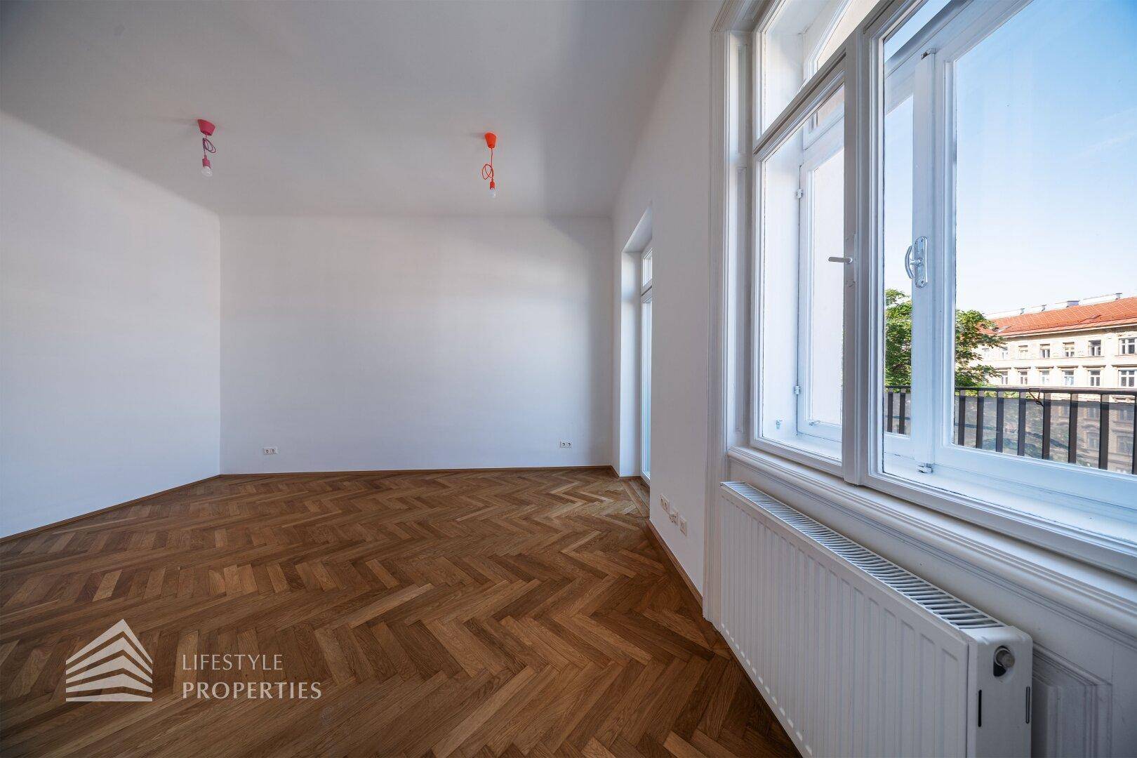 Geräumiger, heller Wohnbereich mit elegantem Fischgrätparkett und großen Fenstern, die viel Tageslicht hereinlassen.