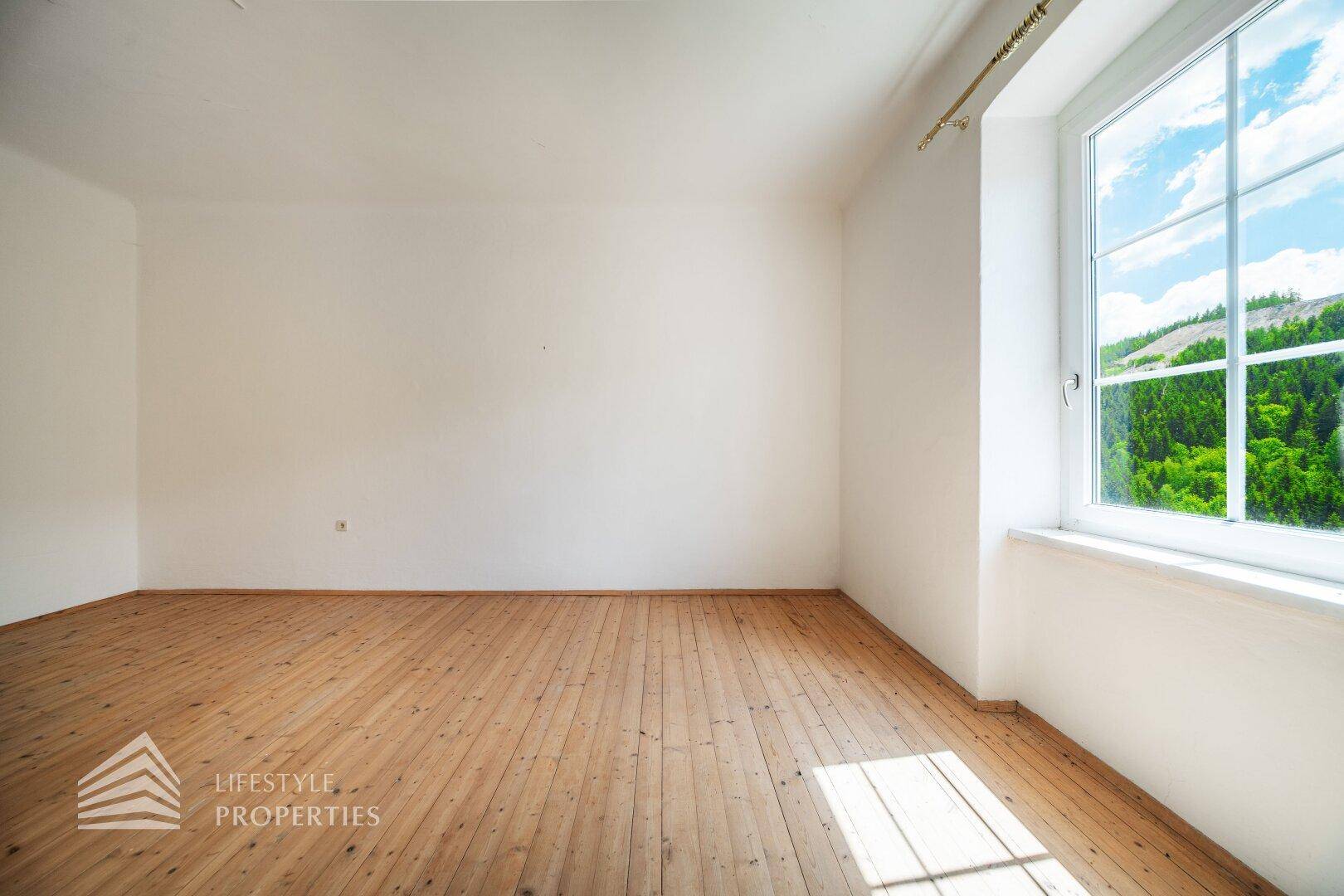 Geräumiges Wohnzimmer mit Holzdielenboden und großem Fenster, das viel Tageslicht hereinlässt.