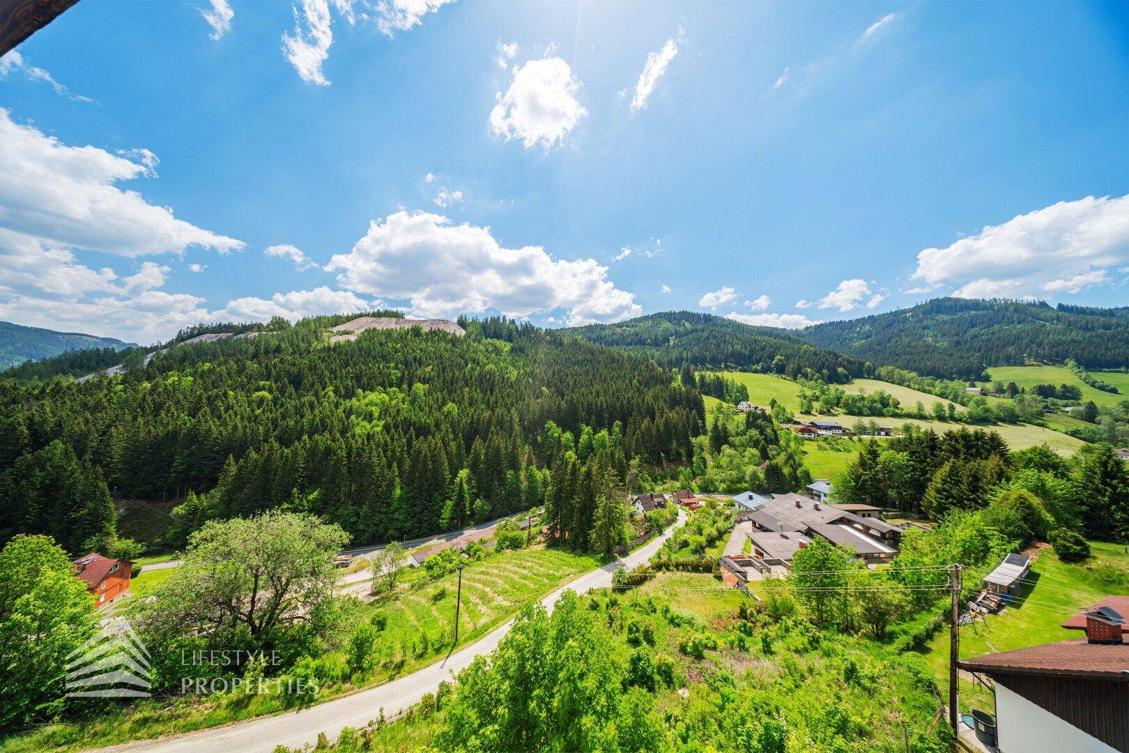 Panoramablick auf die bewaldeten Berge und das Tal mit kleinen Häusern unter blauem Himmel.
