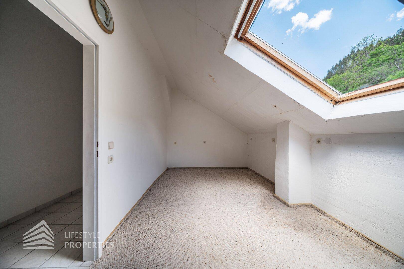 Dachbodenraum mit Teppichboden und Dachschräge, beleuchtet durch ein großes Dachfenster.