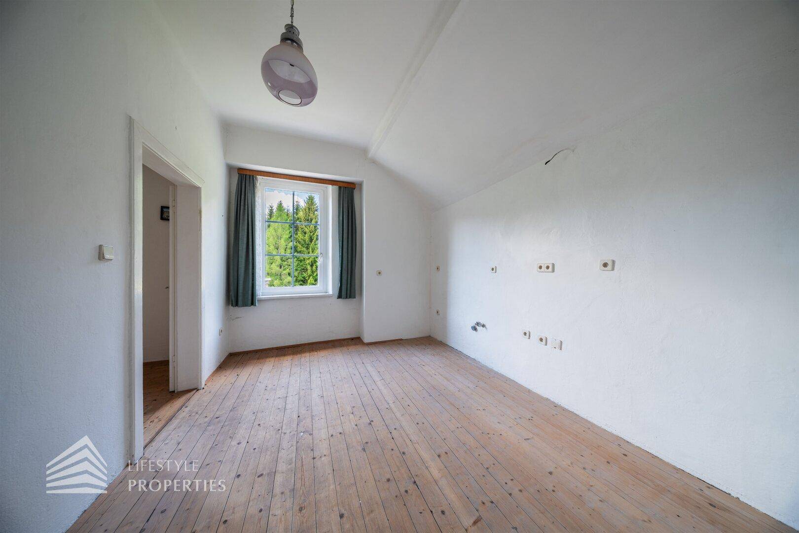 Helles Schlafzimmer mit Holzdielenboden und einem Fenster mit Blick ins Grüne.