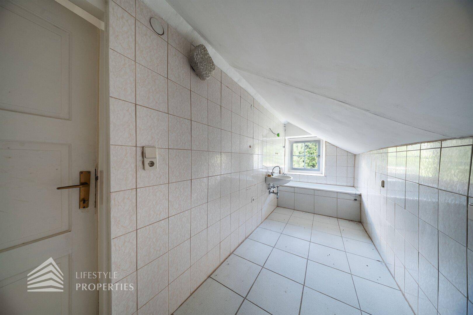 Gefliestes Badezimmer im Dachgeschoss mit Waschbecken und Dachschräge, bereit zur Renovierung.