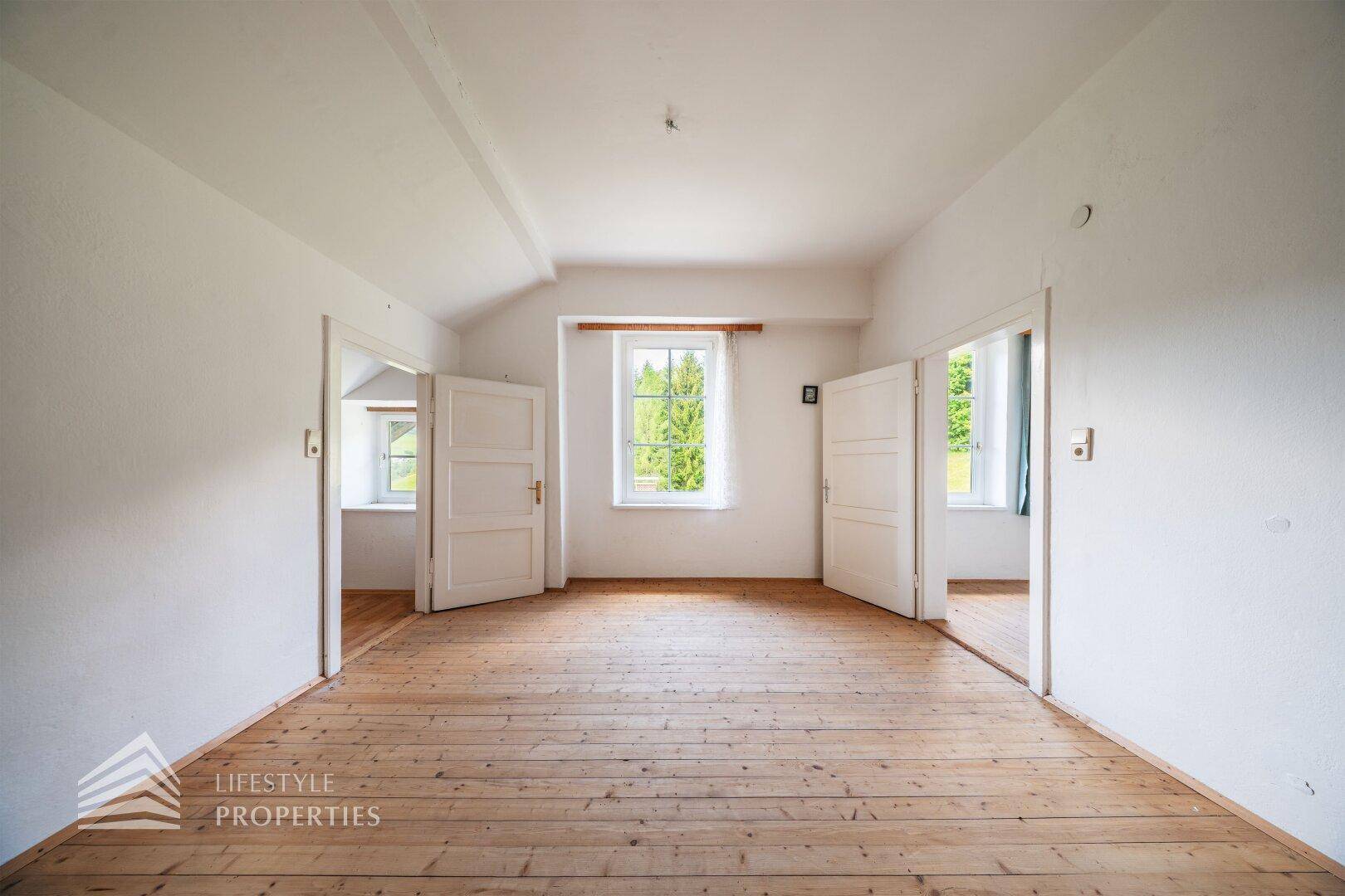 Helles Wohnzimmer mit Holzdielenboden und mehreren Fenstern, die einen Blick ins Grüne bieten.