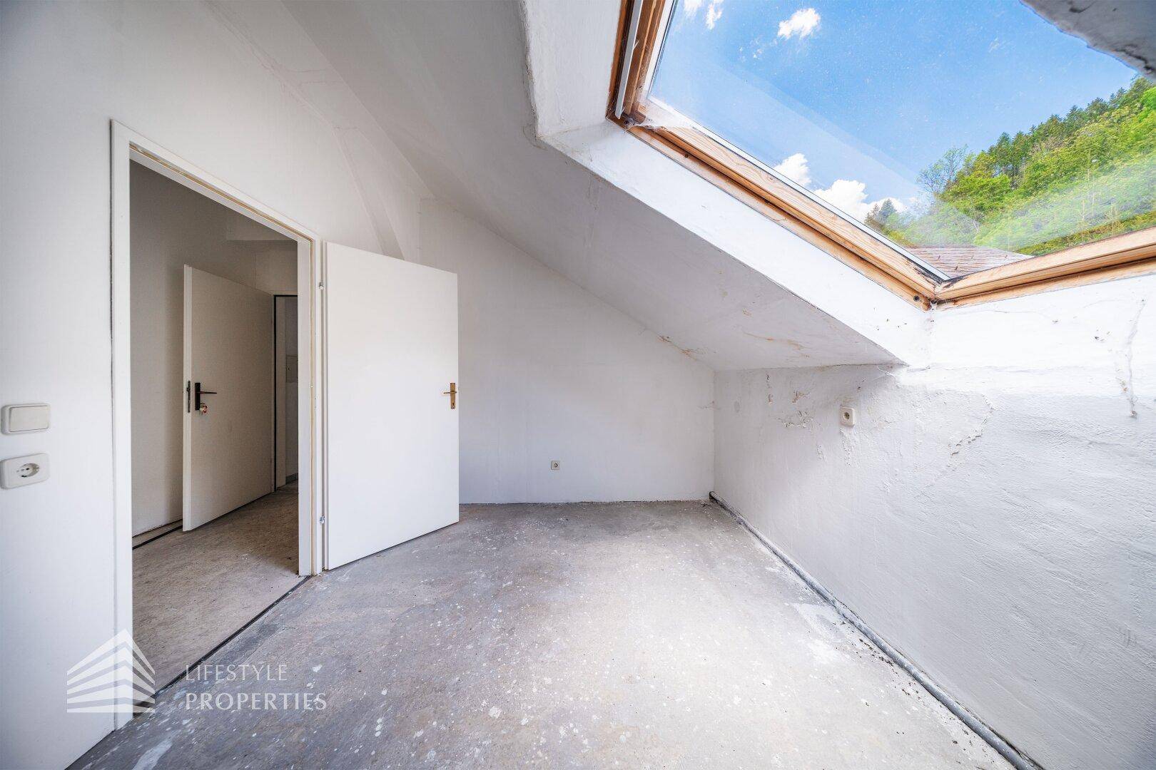 Heller Dachbodenraum mit schrägen Wänden, einem großen Dachfenster und Blick in die Natur.