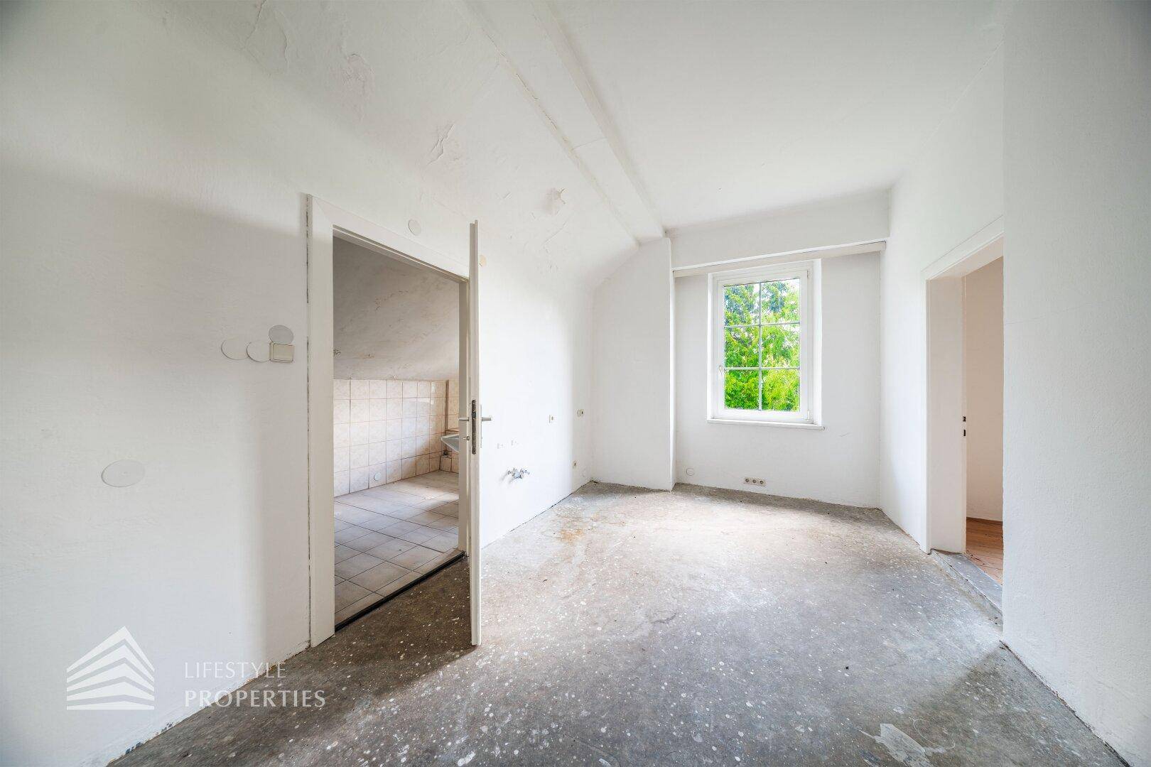 Unausgebauter Dachbodenraum mit Fenster und Blick in ein gefliestes Badezimmer.