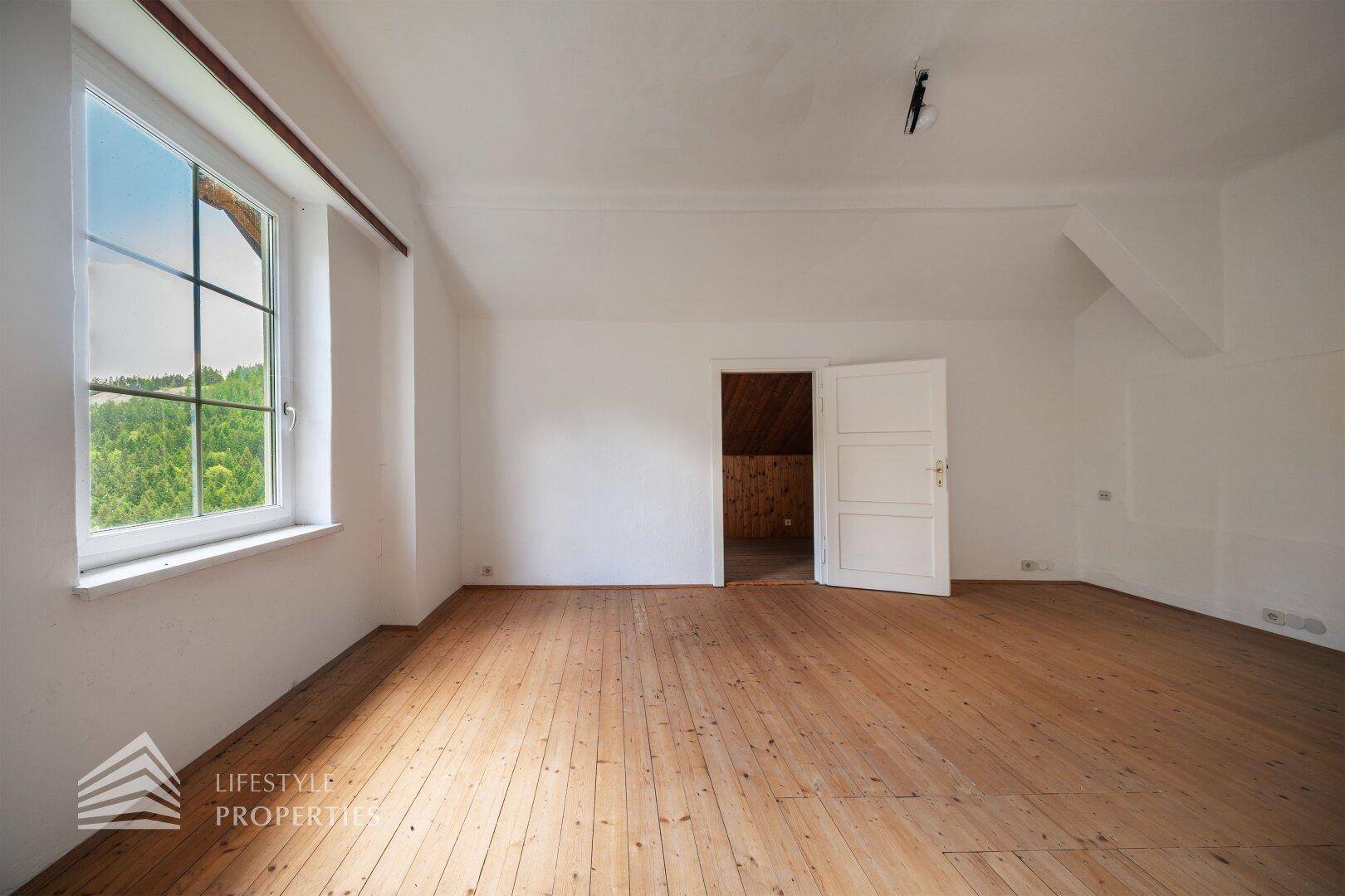 Geräumiges Zimmer mit Holzdielenboden und großem Fenster mit Blick ins Grüne.