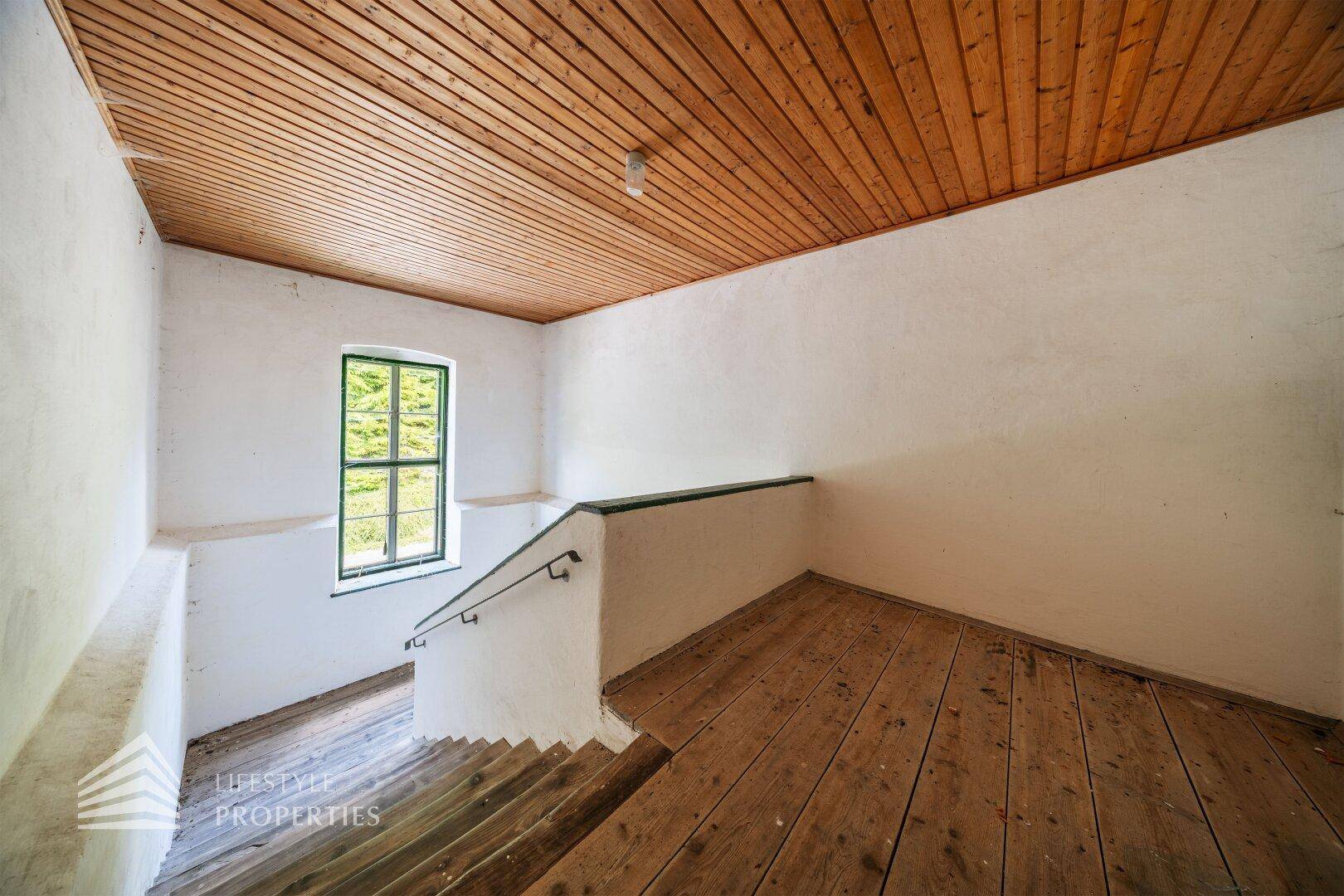 Treppenhaus mit Holzdielenboden, Holzdecke und Fenster mit Blick ins Grüne.