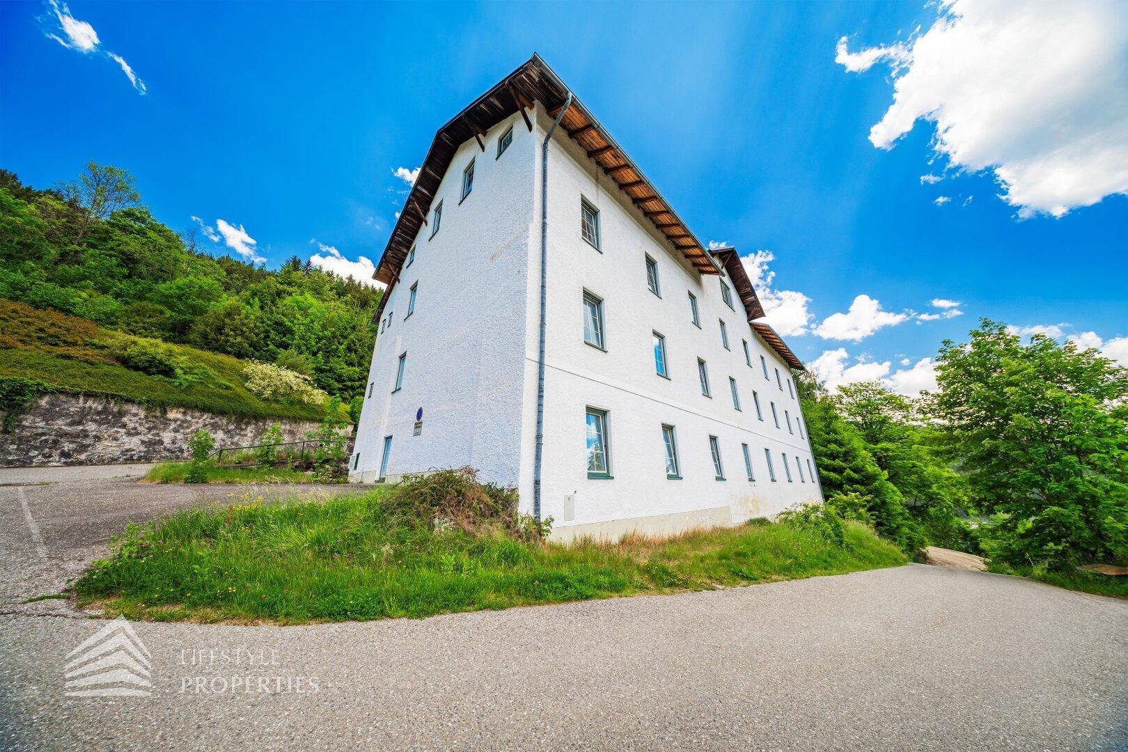 Seitenansicht des weißen Gebäudes mit grünen Akzenten, eingebettet in eine grüne Hügellandschaft unter blauem Himmel.