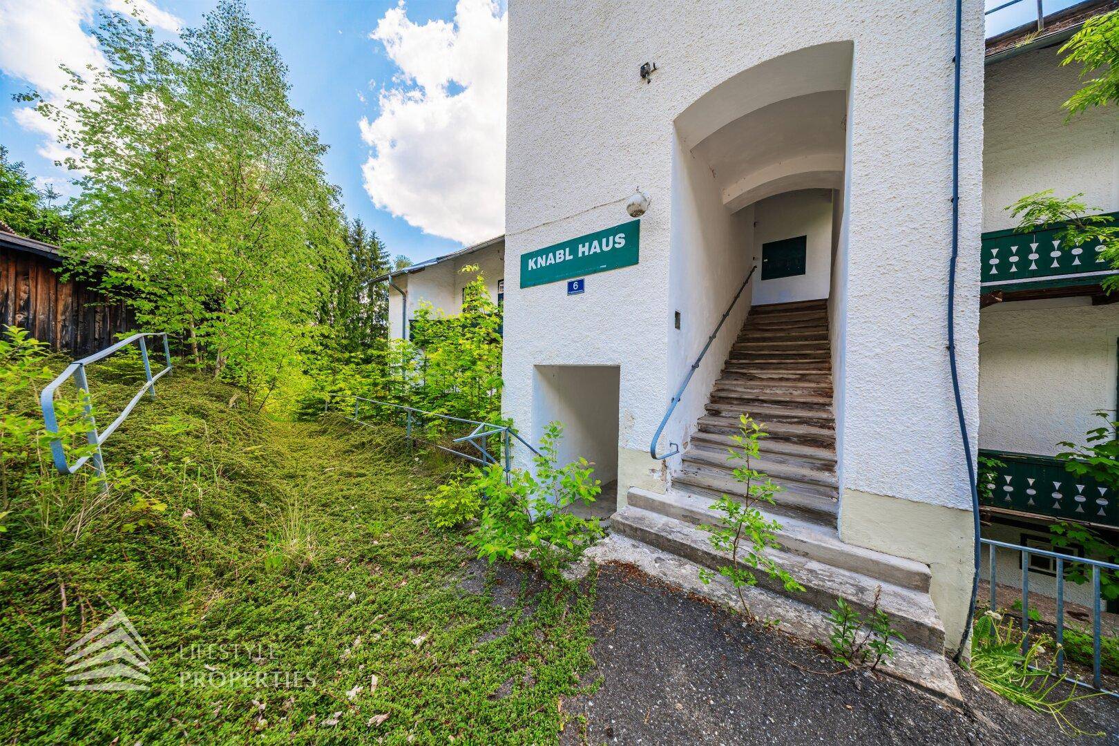 Eingangsbereich des Gebäudes mit Treppe und Geländer, umgeben von wilder Vegetation und Bäumen.