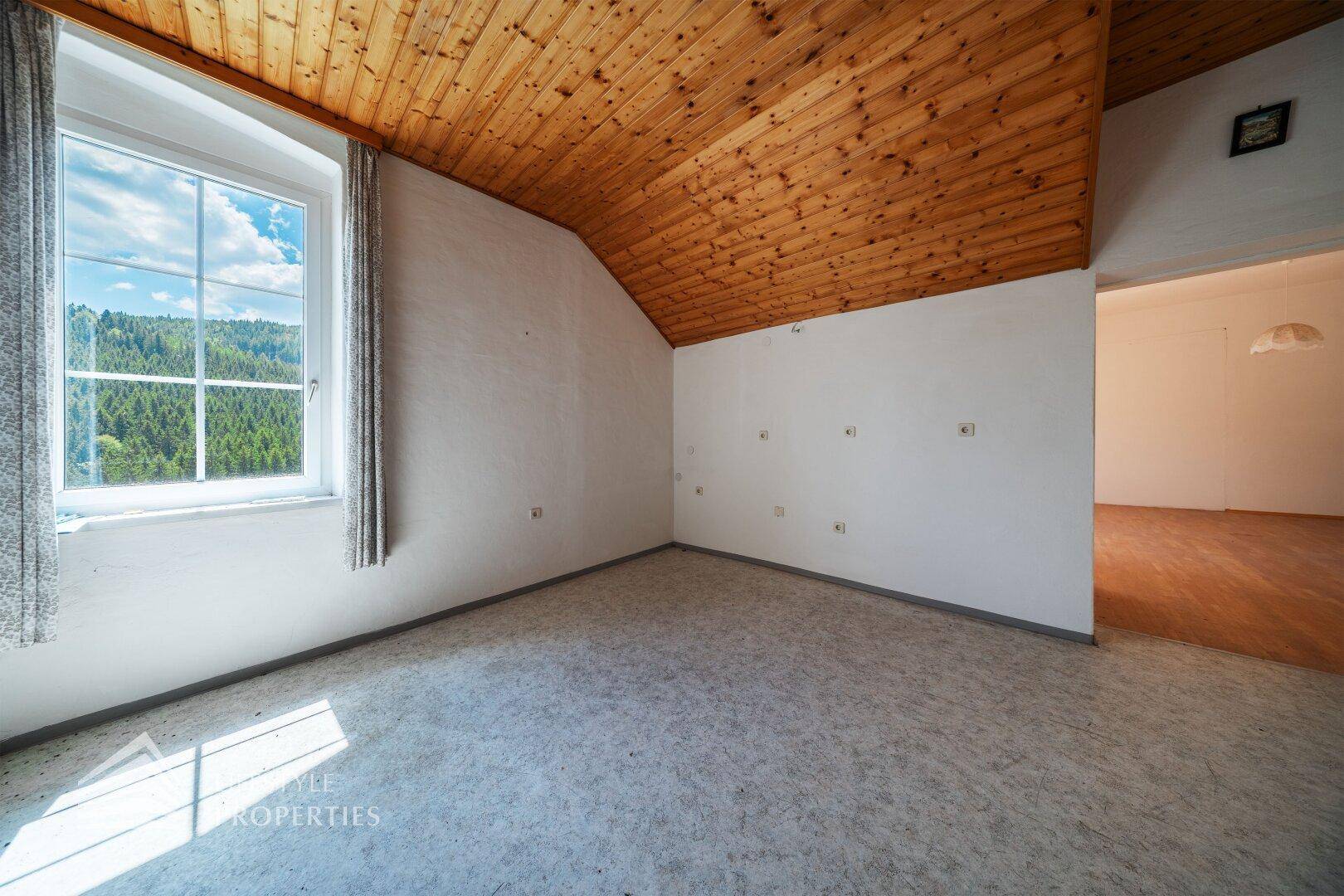 Heller Dachraum mit Holzdecke, Teppichboden und weitem Blick in die grüne Landschaft.