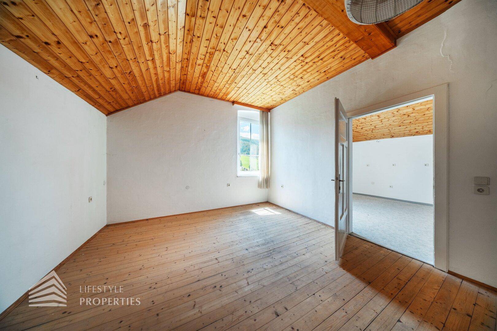 Helles Zimmer im Dachgeschoss mit Holzdecke, Holzdielenboden und Fenster mit Blick ins Grüne.