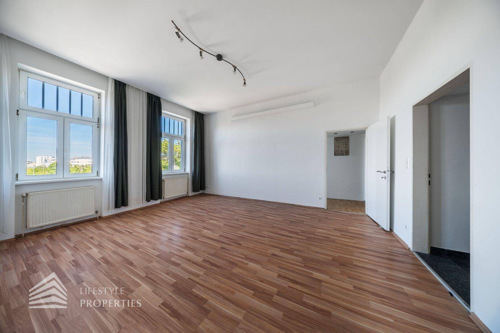 Geräumiges, helles Zimmer mit Holzboden und Blick nach draußen durch große Fenster.