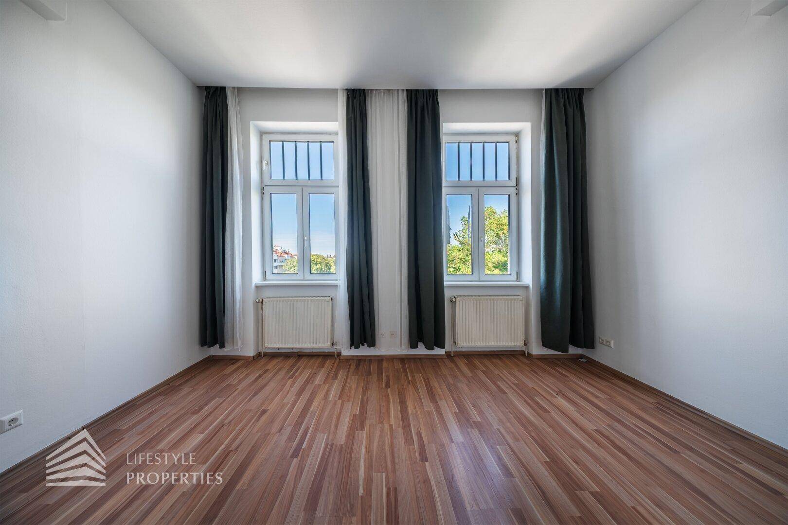 Leeres Zimmer mit zwei hohen Fenstern, die viel Tageslicht hereinlassen und einen Ausblick bieten.