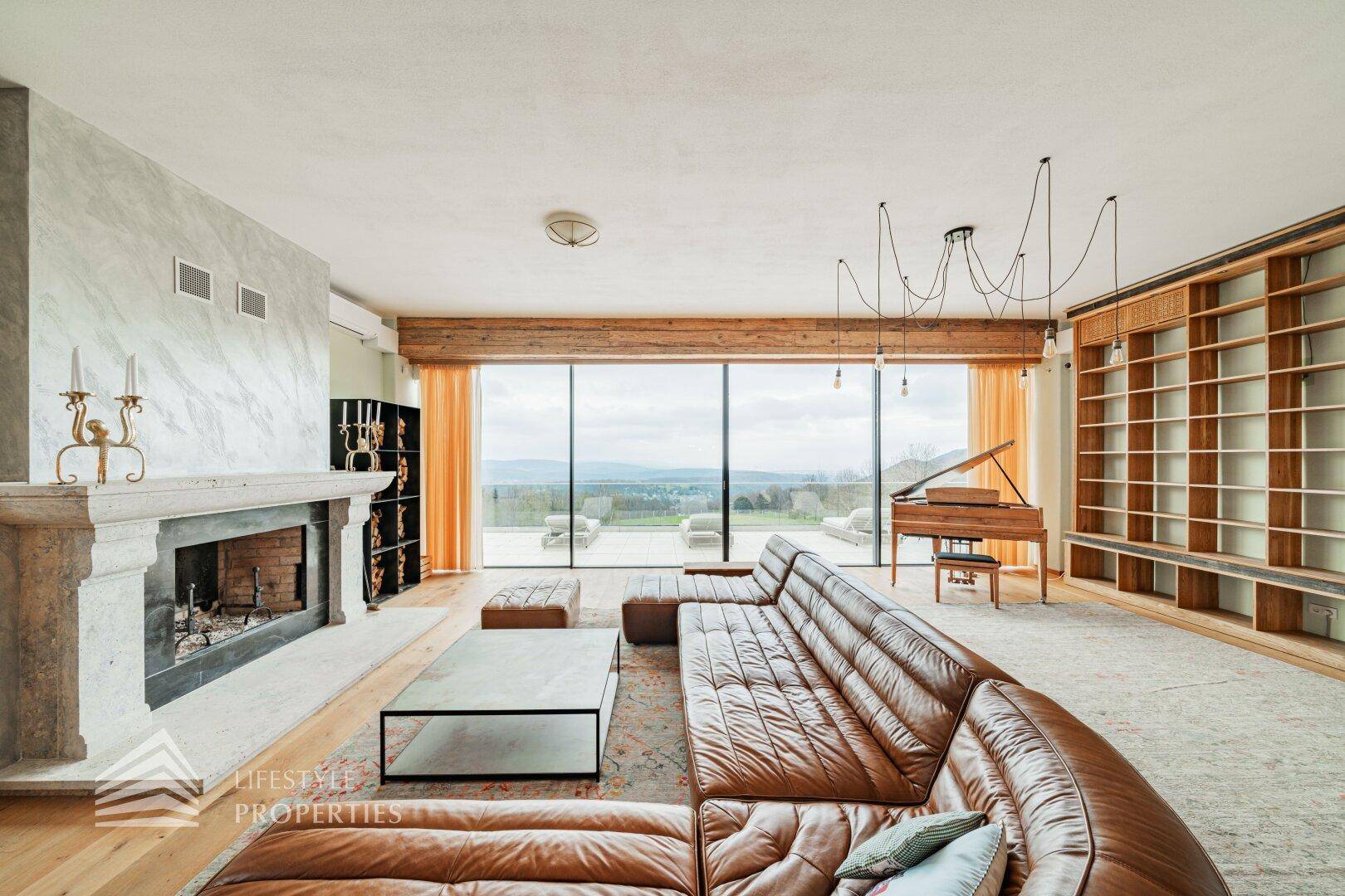 Helles Wohnzimmer mit Ledersofa, Kamin und raumhohen Fenstern, die einen weiten Blick bieten.