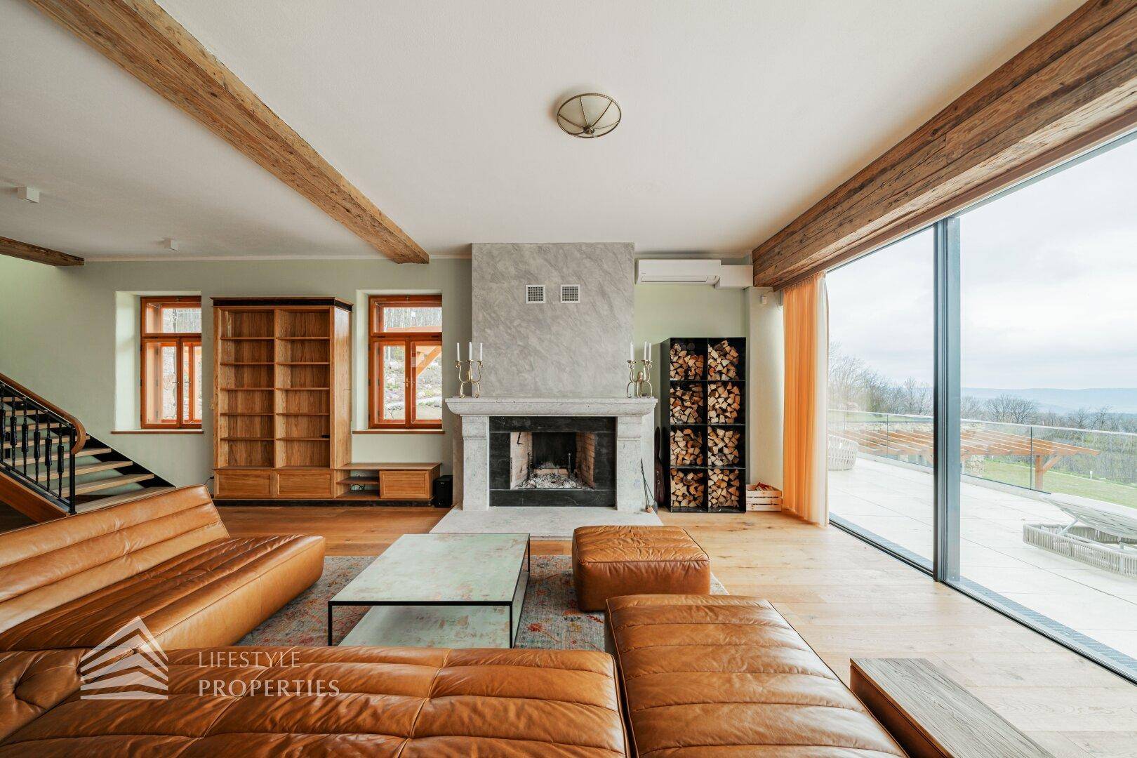 Gemütliches Wohnzimmer mit Kamin, Ledersofa und großen Fenstern mit Blick auf die Natur.