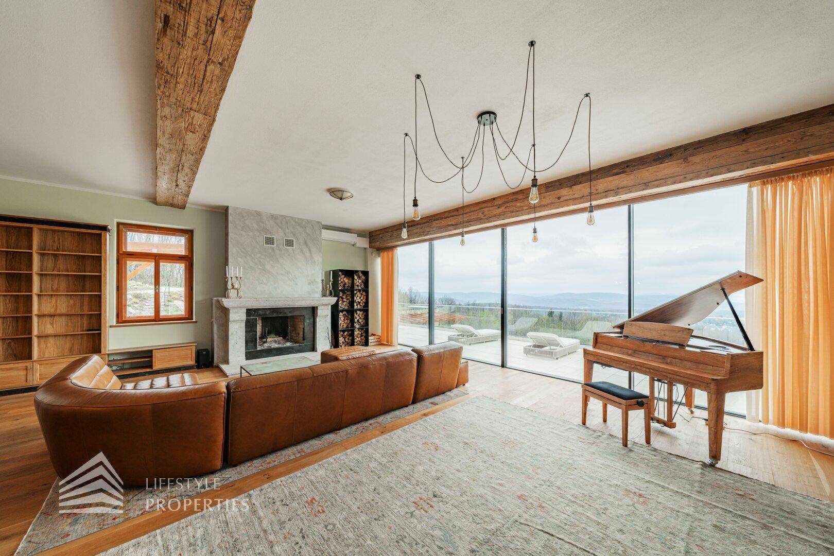 Geräumiges Wohnzimmer mit Ledersofa, Flügel, Kamin und Panoramafenstern mit weitem Ausblick.