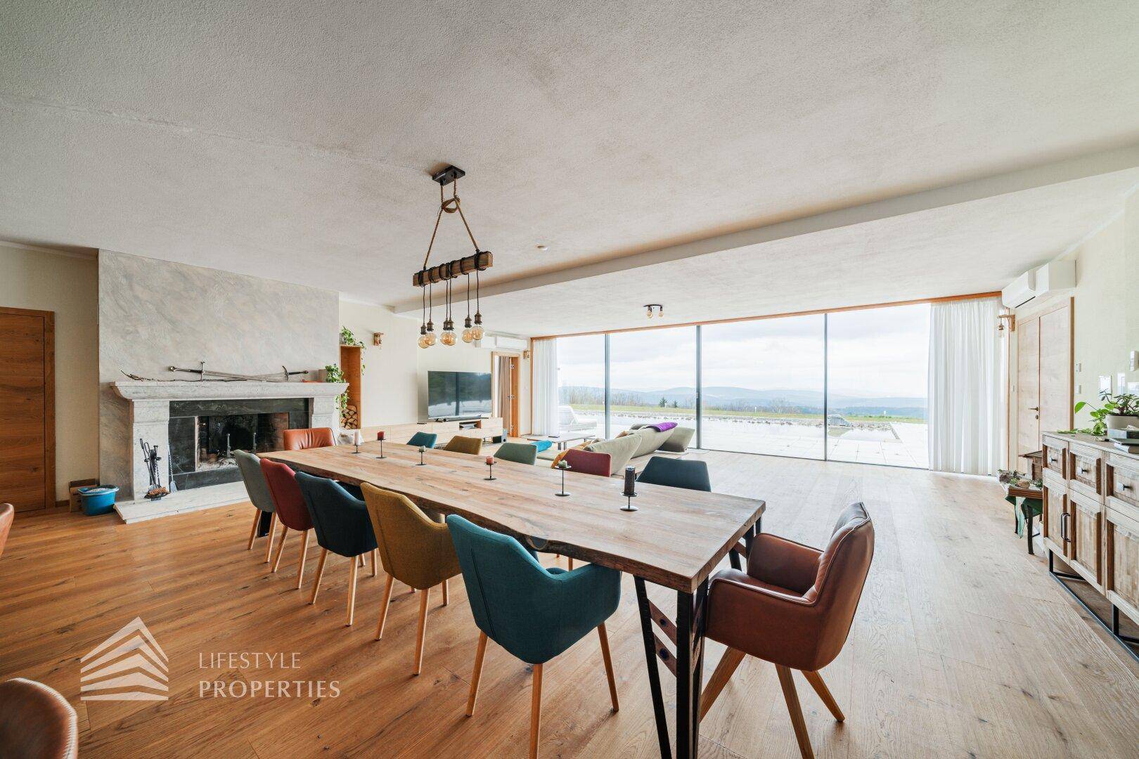 Geräumiger Essbereich mit Holztisch, bunten Stühlen und Kamin, offen zum Wohnbereich mit Weitblick.
