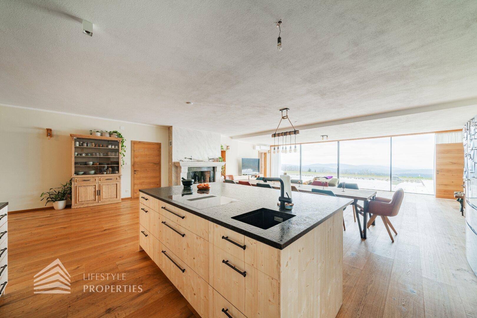 Moderne Holzküche mit Insel und dunkler Arbeitsplatte, offen zum Ess- und Wohnbereich mit Weitblick.