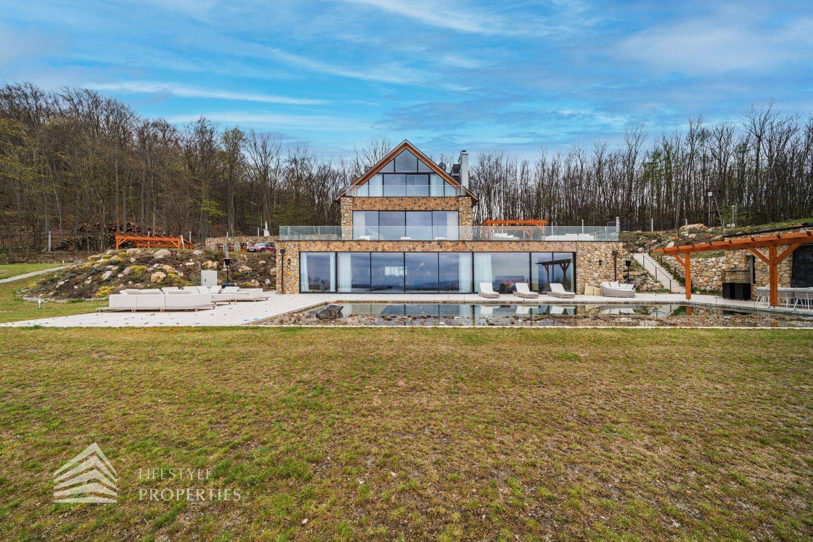 Moderne Villa mit großen Glasfronten, Infinity-Pool und weitläufigem Rasenbereich vor bewaldetem Hintergrund.