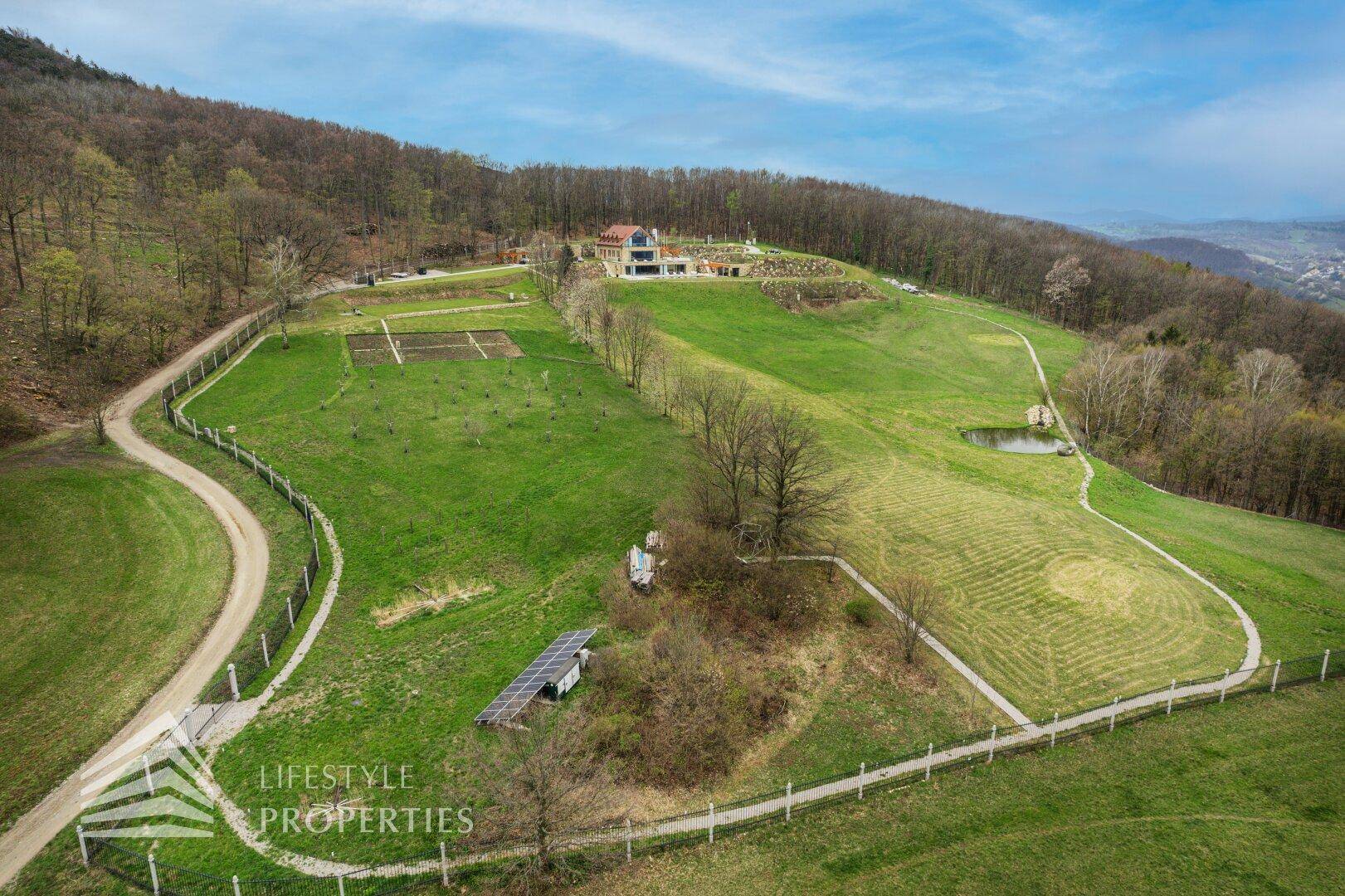 Weitläufiges Grundstück mit grünen Hügeln, Waldrand und einem kleinen Teich in ländlicher Umgebung.