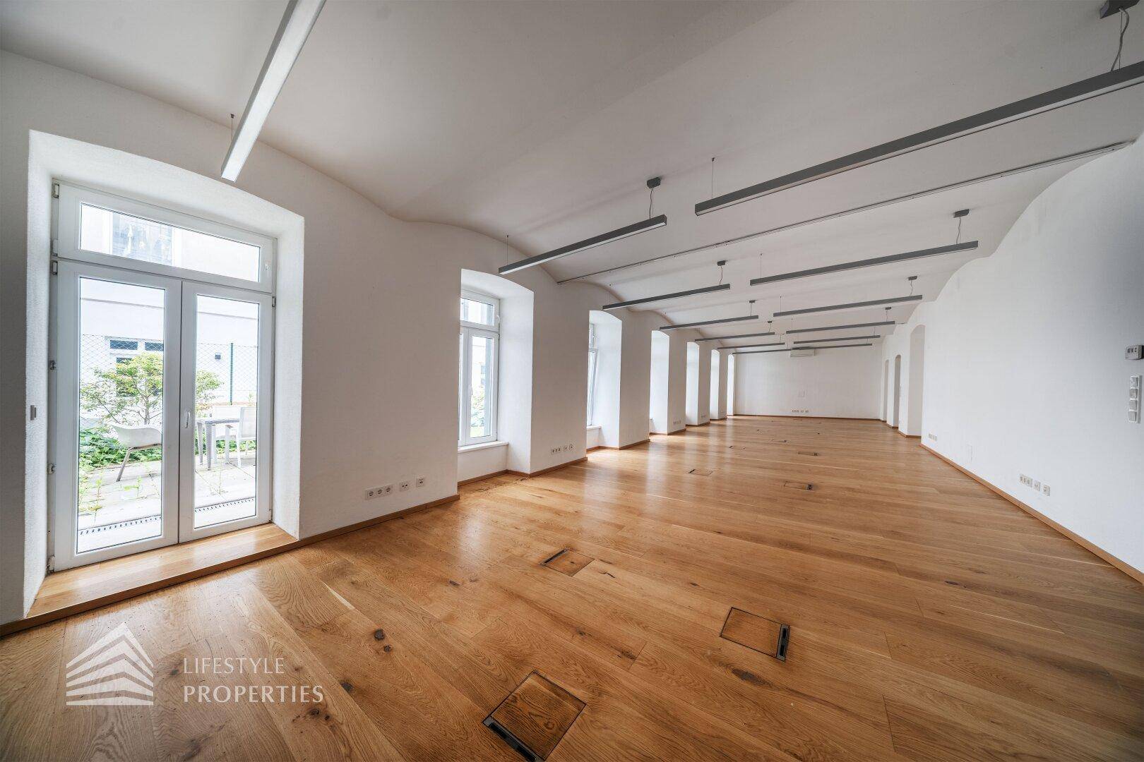 Geräumiger Büroraum mit Holzdielenboden, hohen Fenstern und Zugang zu einem Außenbereich.
