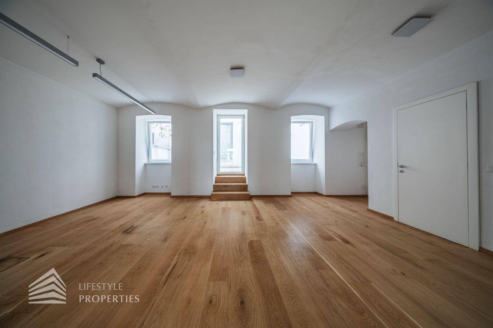 Heller, offener Raum mit Holzdielenboden und gewölbten Fenstern, die über Holzstufen zugänglich sind.