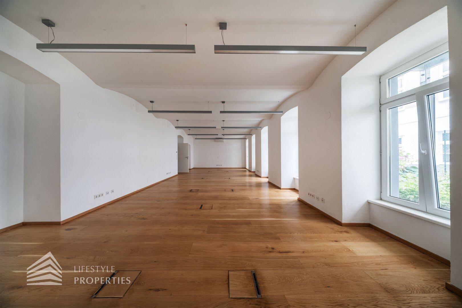 Langer, heller Raum mit Holzboden, hohen Fenstern und modernen Deckenleuchten.