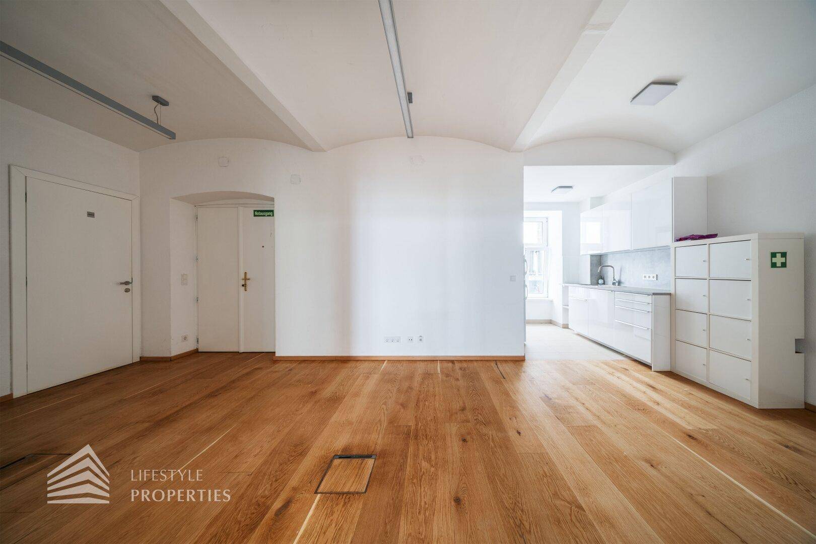 Großer Büroraum mit Holzboden, weißen Wänden und angrenzender, moderner Küchenzeile.