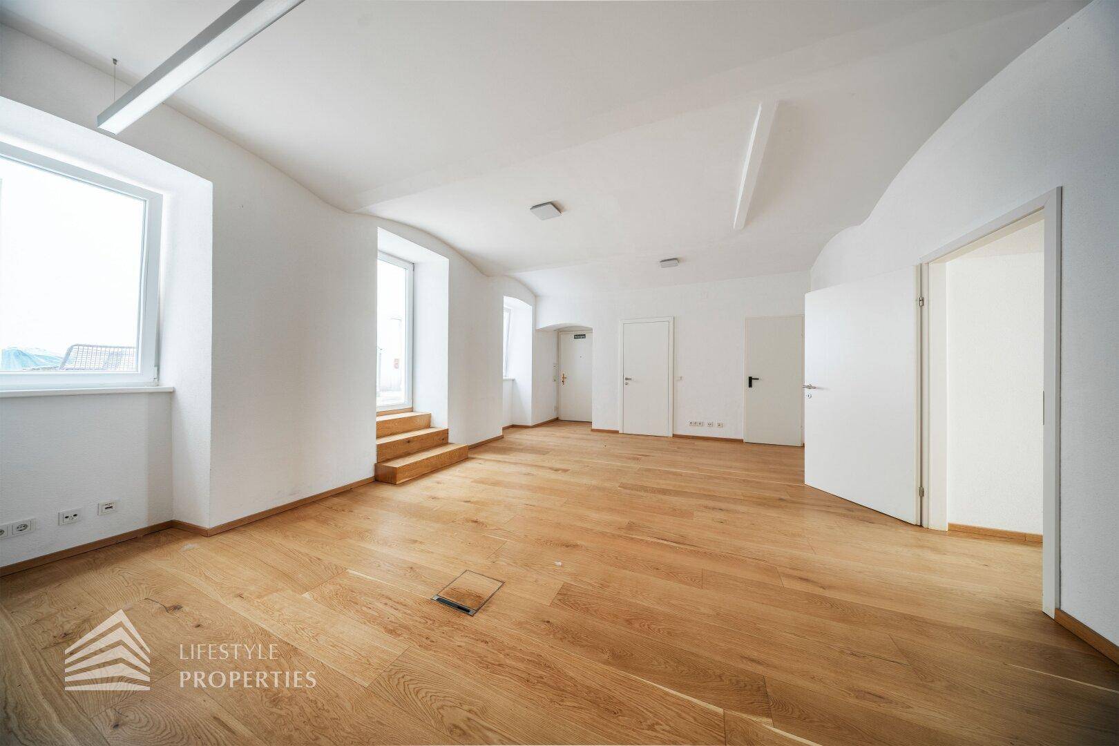 Geräumiger Raum mit Holzboden, weißen Wänden und Fenstern, die über kleine Stufen erreichbar sind.