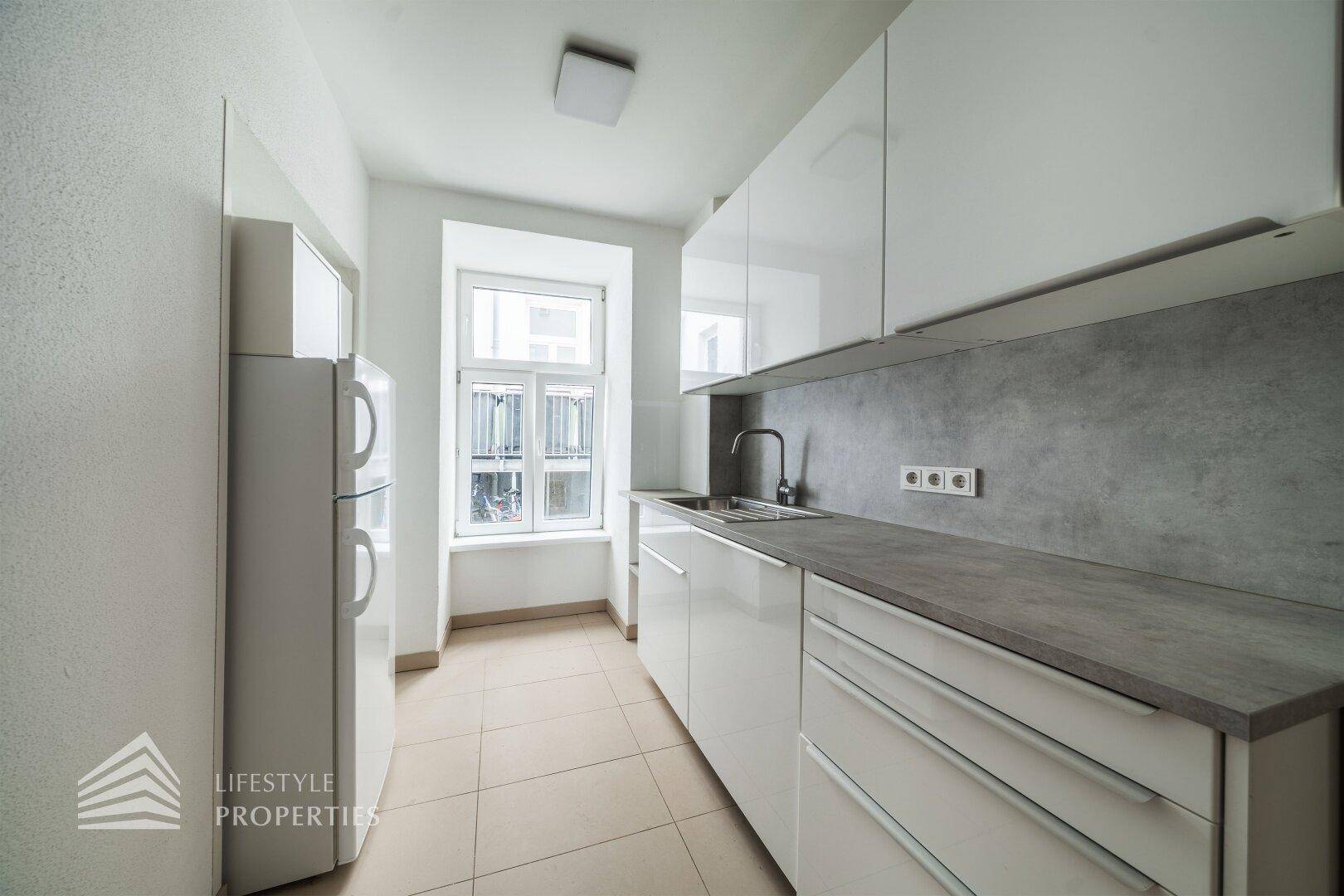 Moderne Küchenzeile mit weißen Hochglanzschränken, grauer Arbeitsplatte und Fenster.