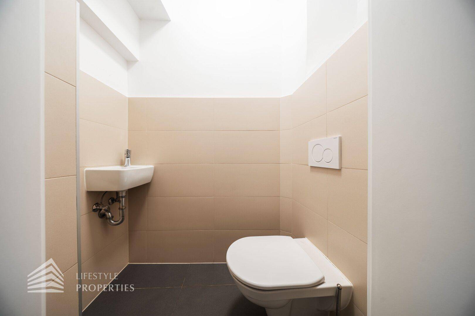 Kleines Gäste-WC mit beigefarbenen Fliesen, dunklem Boden und wandhängendem WC.