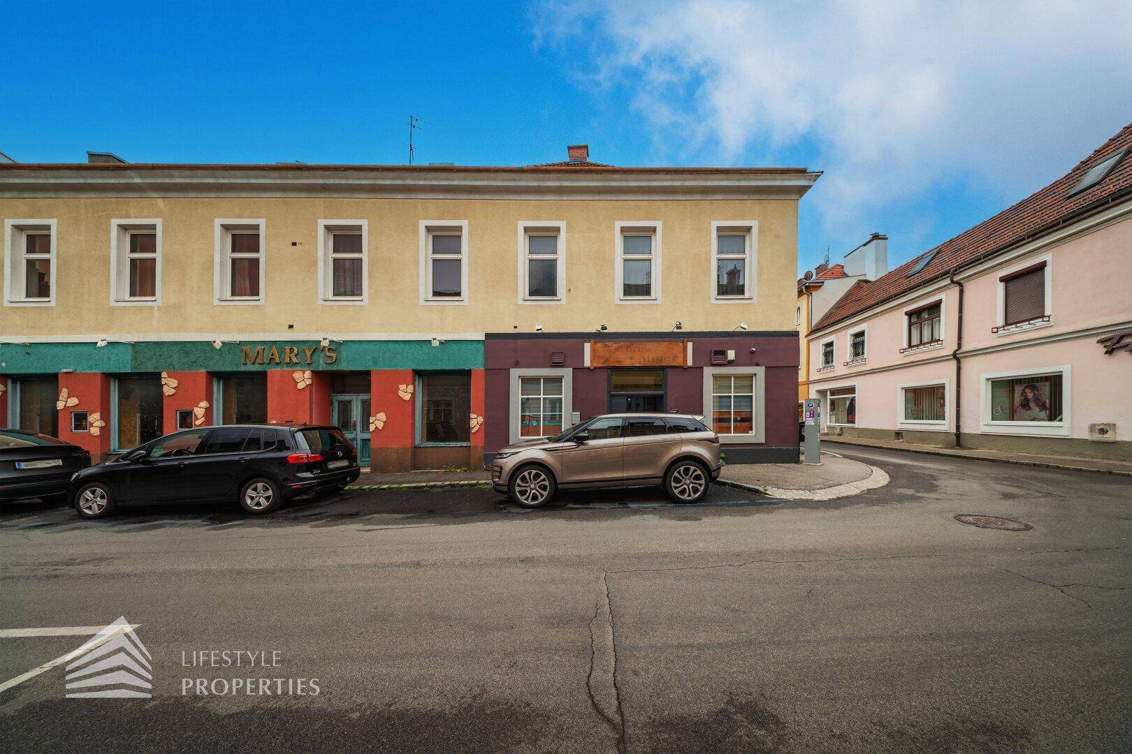 Fassade eines Gebäudes mit Geschäften im Erdgeschoss und geparkten Fahrzeugen auf der Straße.